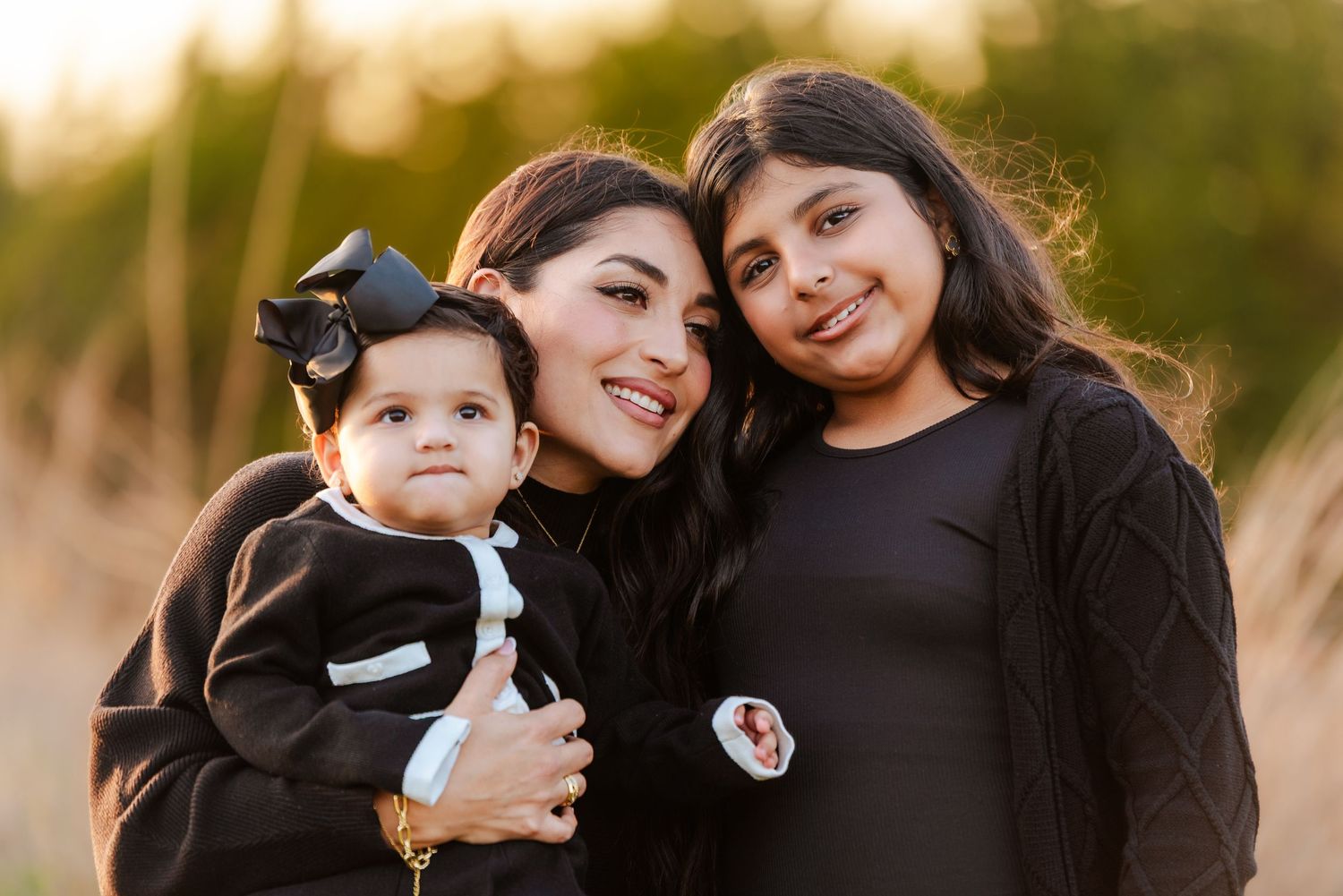 A mother smiles warmly while holding her baby and posing with her young daughter in a sunny outdoor setting.
