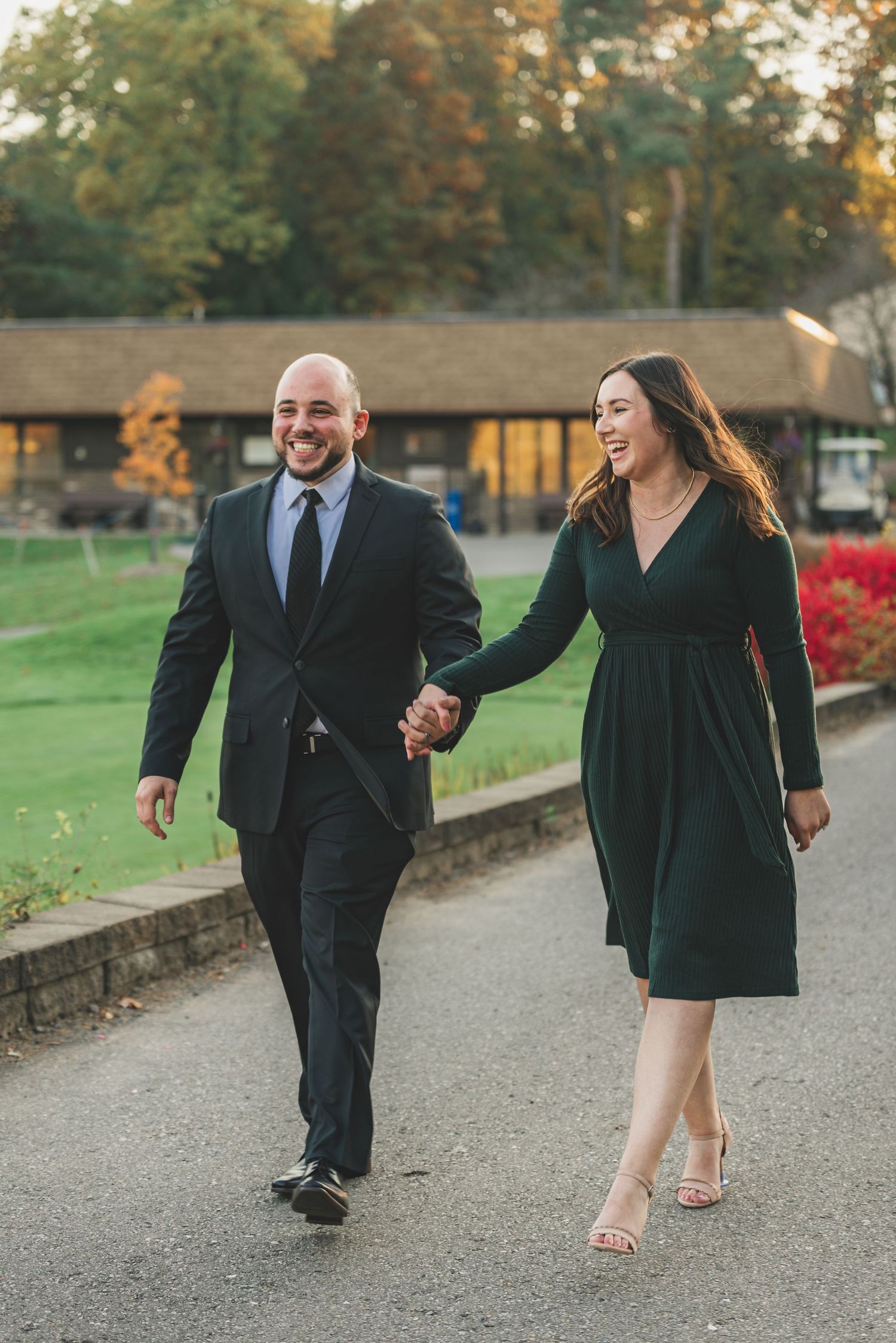 Smiling couple holding hands walking along path during fall engagement photo session outdoors.