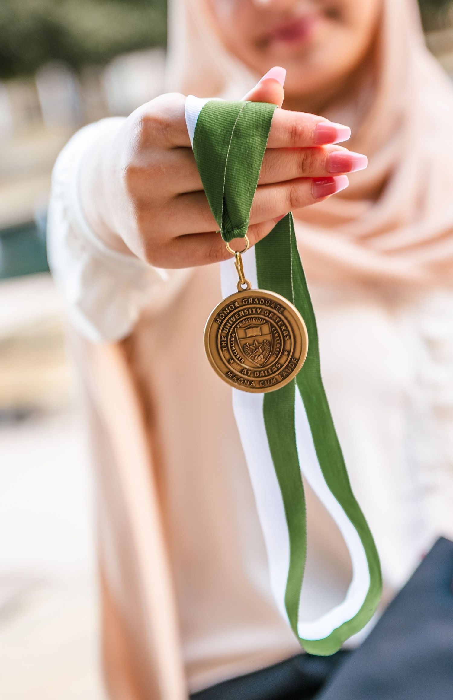 A woman in a hijab proudly displays a bronze university medal on a green ribbon.