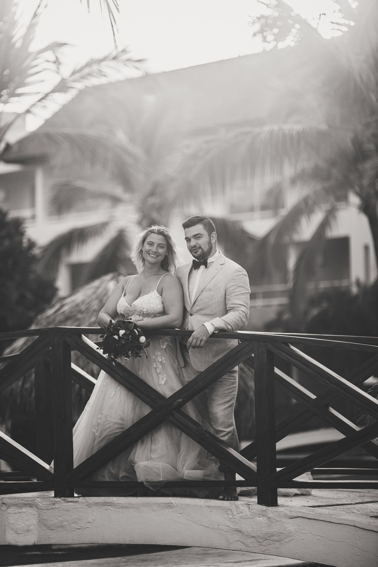 Bride and groom pose on wooden bridge in elegant black and white tropical resort wedding photo.