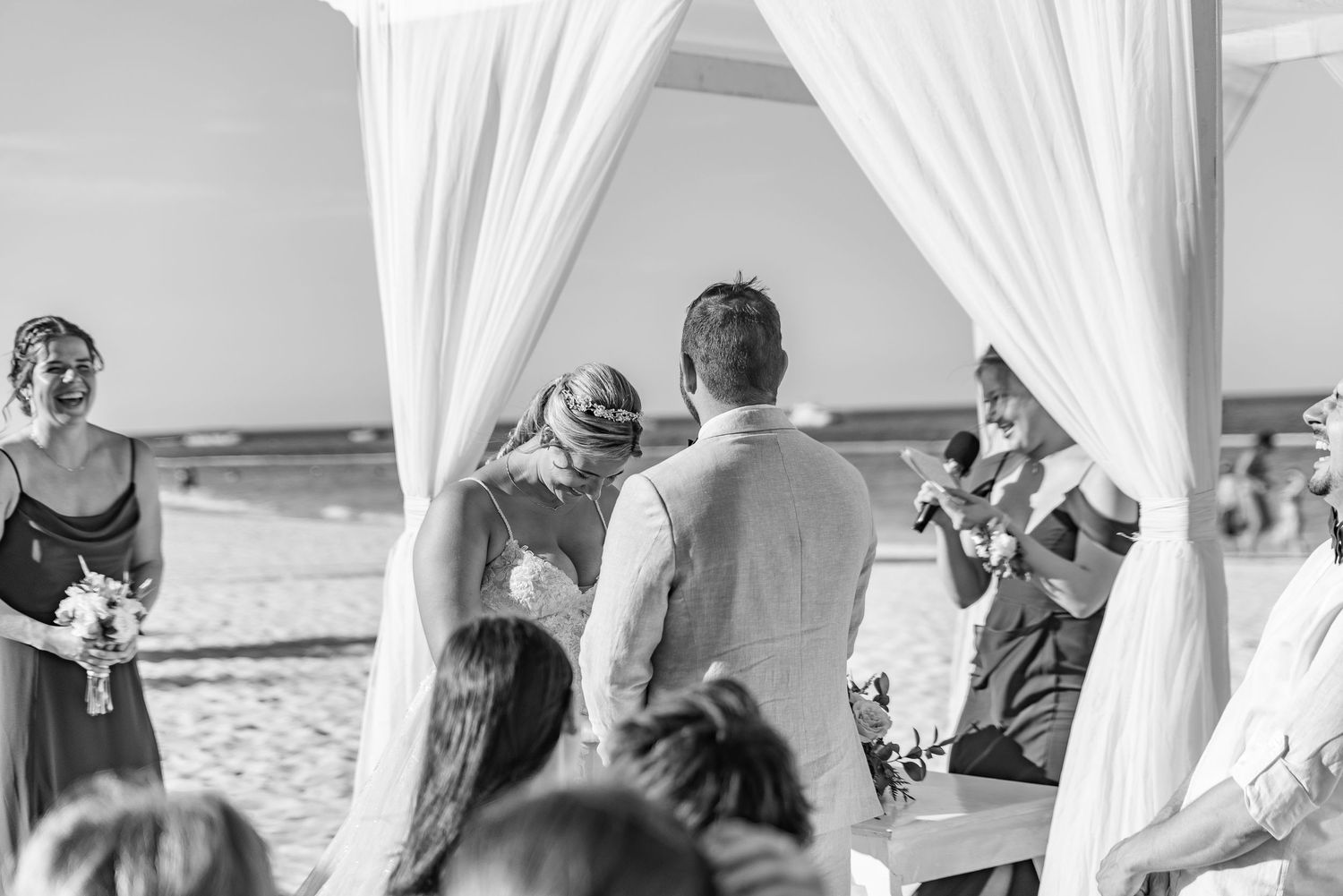 Black and white beach wedding ceremony with couple exchanging vows under elegant white draped arch.