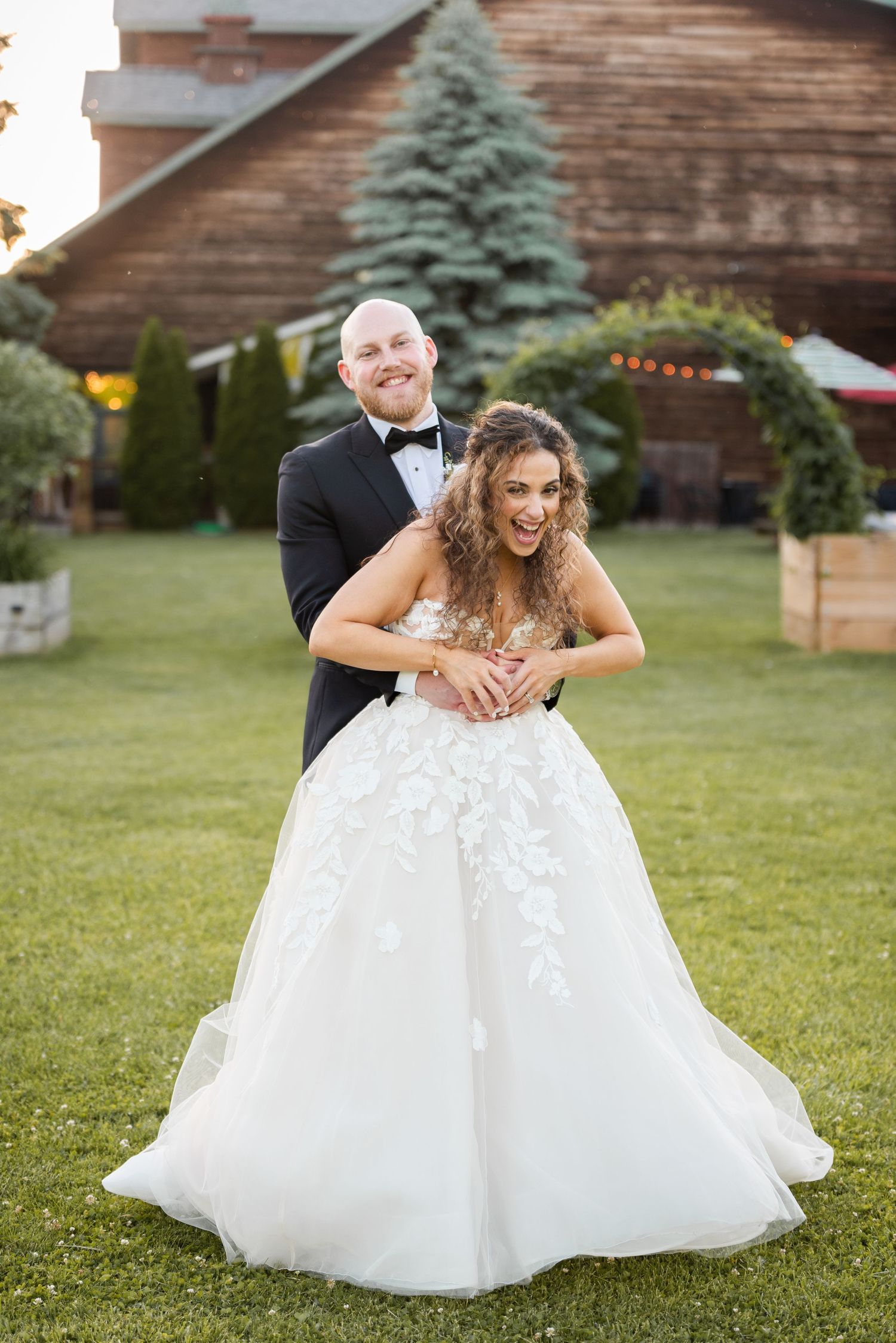 Joyful bride in floral ballgown embraced by groom in tuxedo on green lawn at barn wedding.