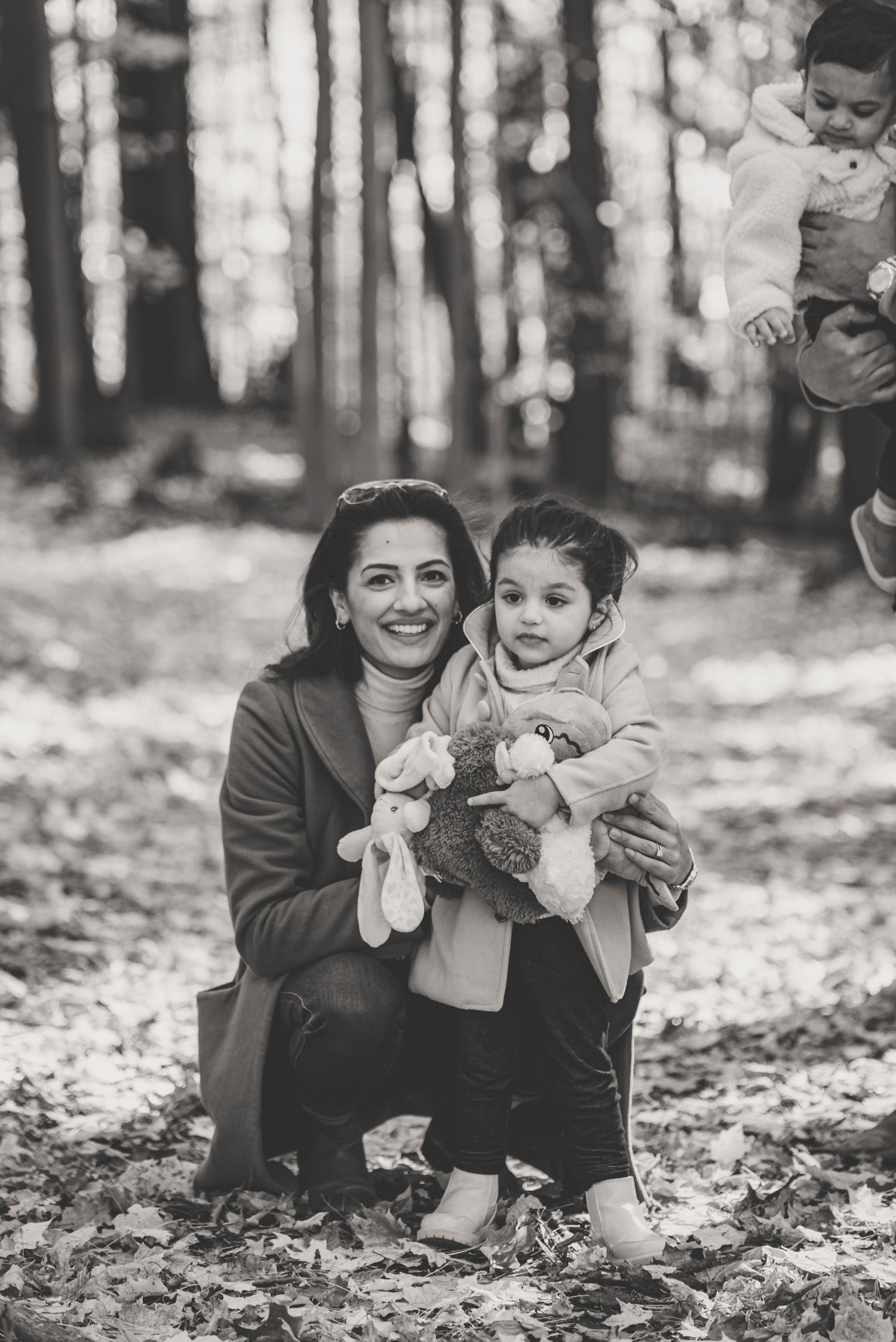 Smiling mother kneeling with young daughter holding stuffed animals in autumn forest in black and white.