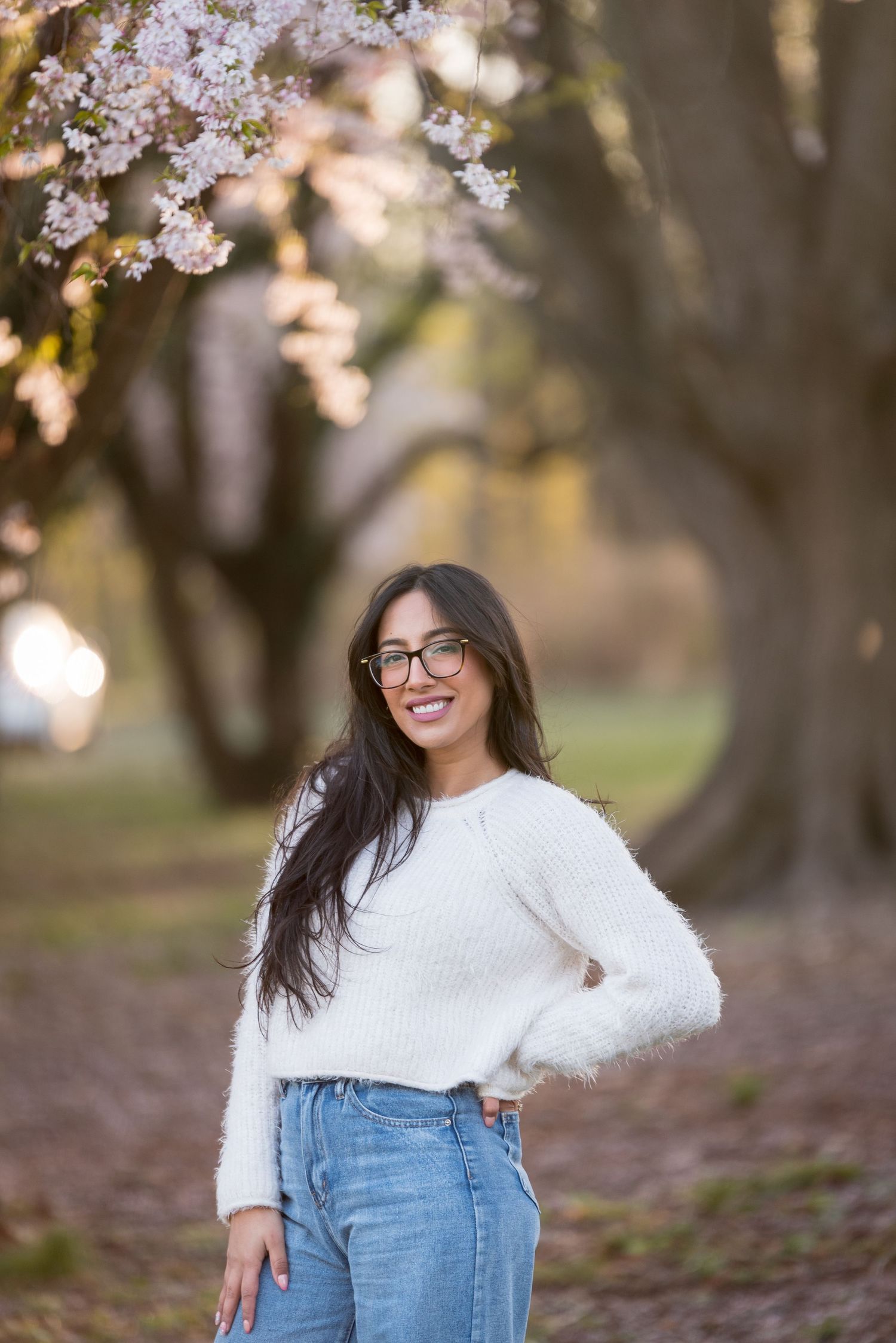 Young woman in white sweater and jeans smiling under blooming cherry blossom trees in spring.