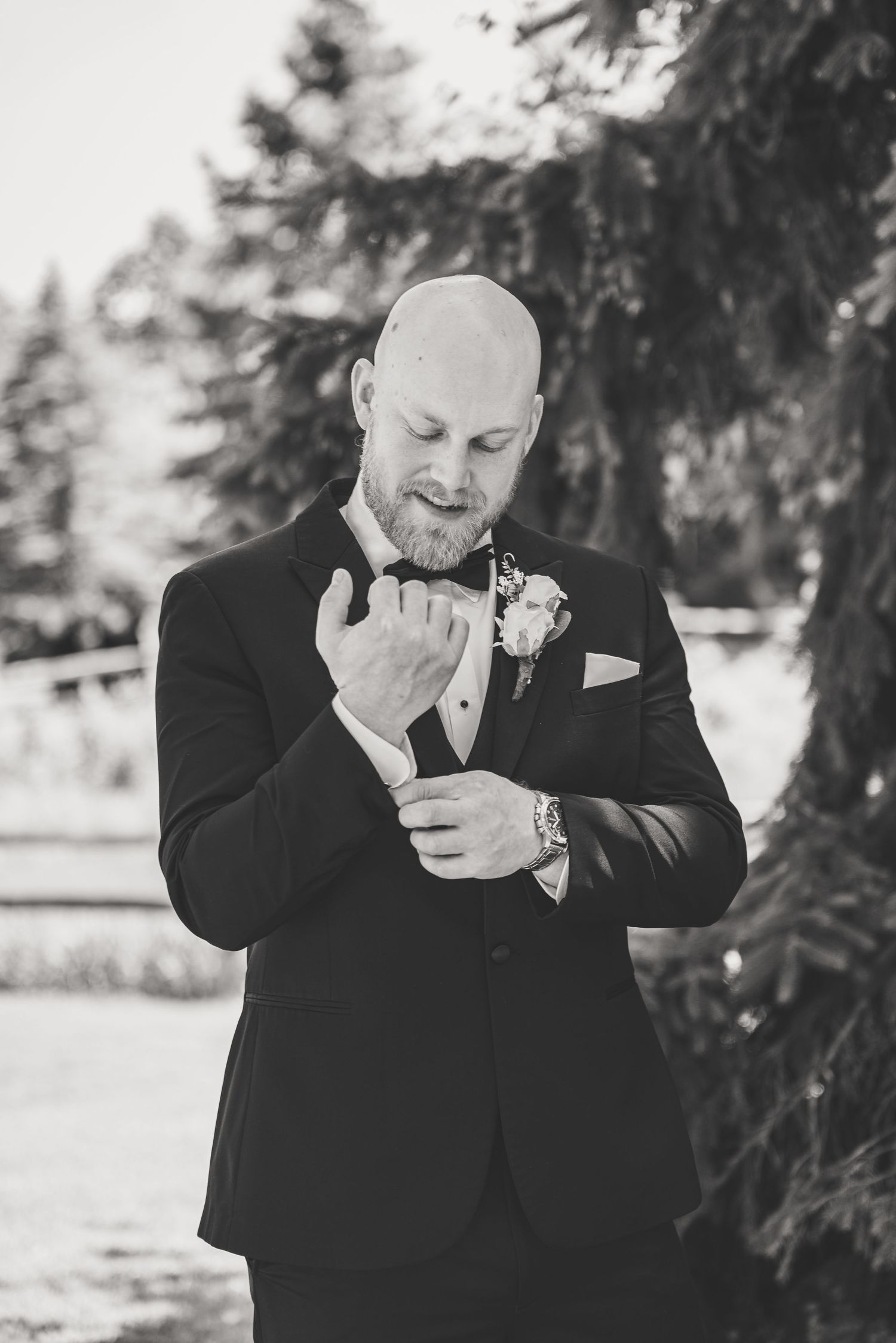 Groom in black tuxedo adjusting cufflinks outdoors in black and white photo.
