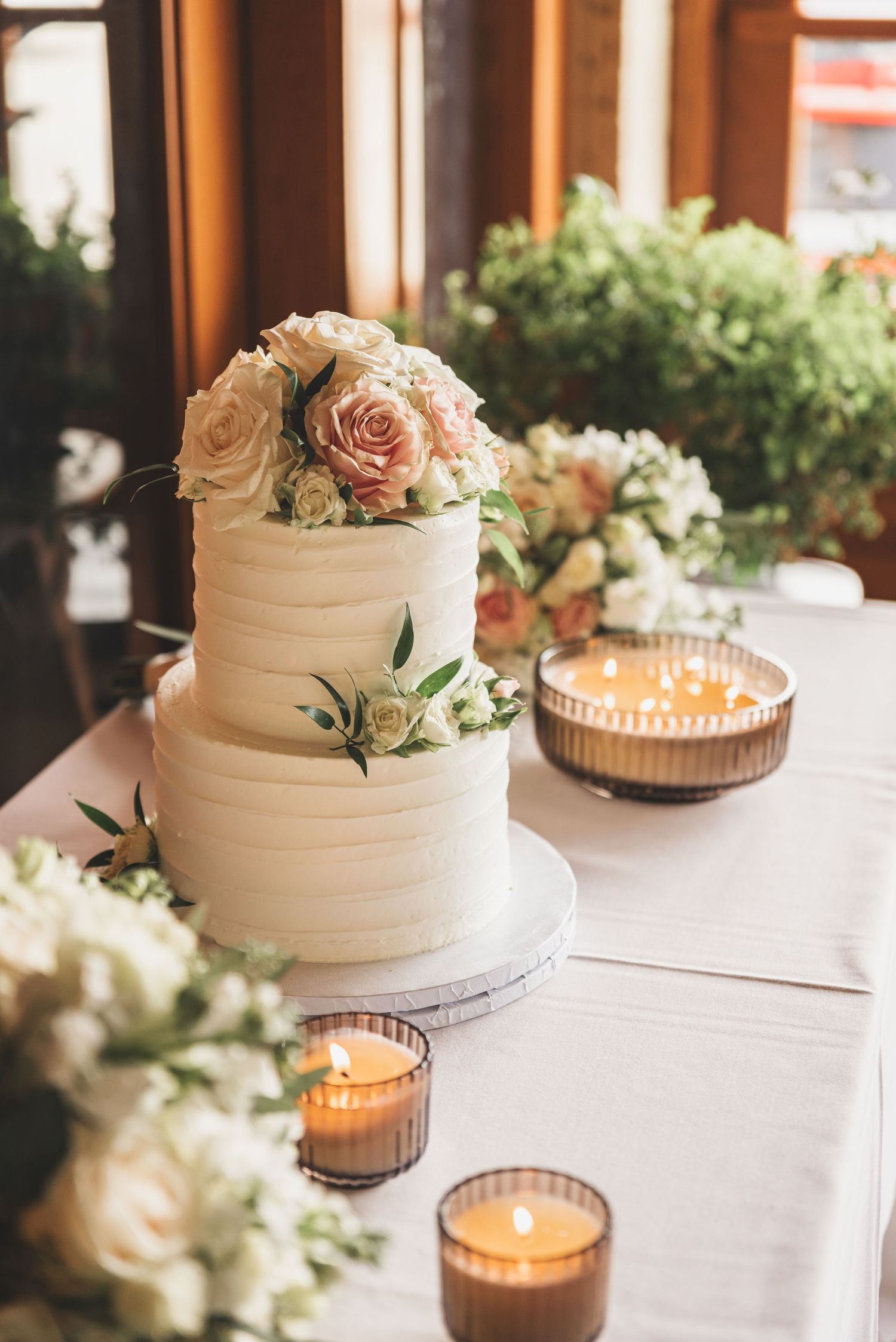 Elegant two-tier white wedding cake topped with pink roses surrounded by candles and floral arrangements.