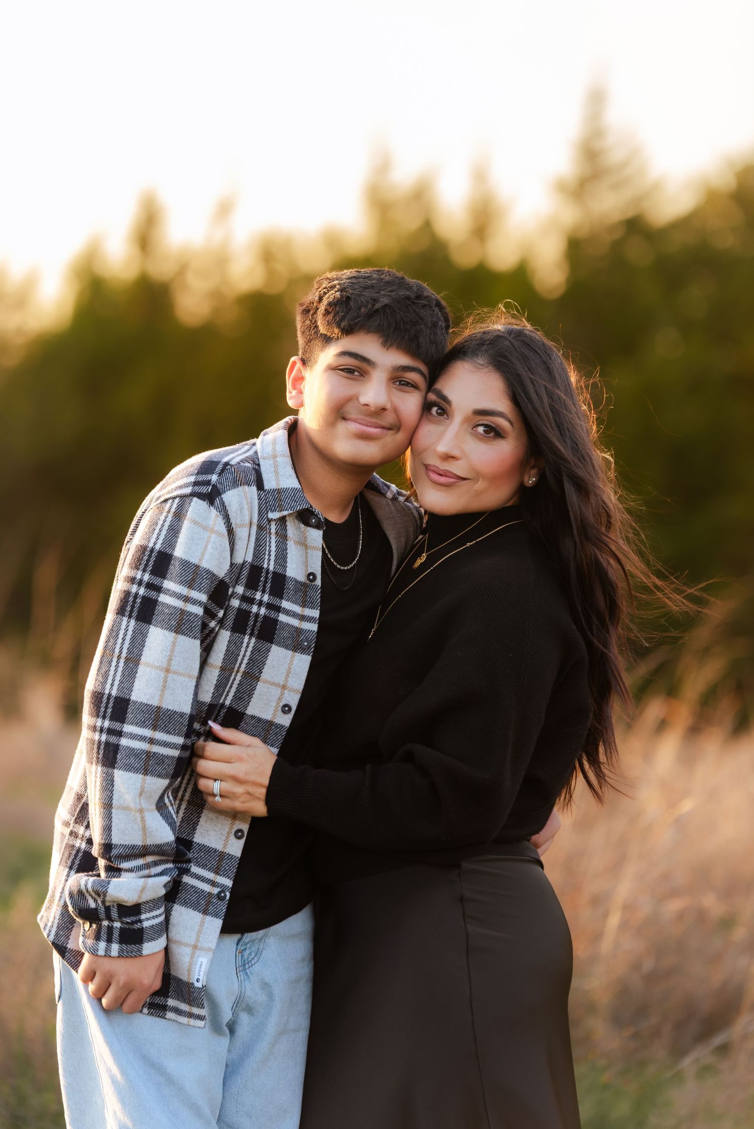 A teenage boy and woman embrace warmly in a sunlit field during a golden hour family photo session.