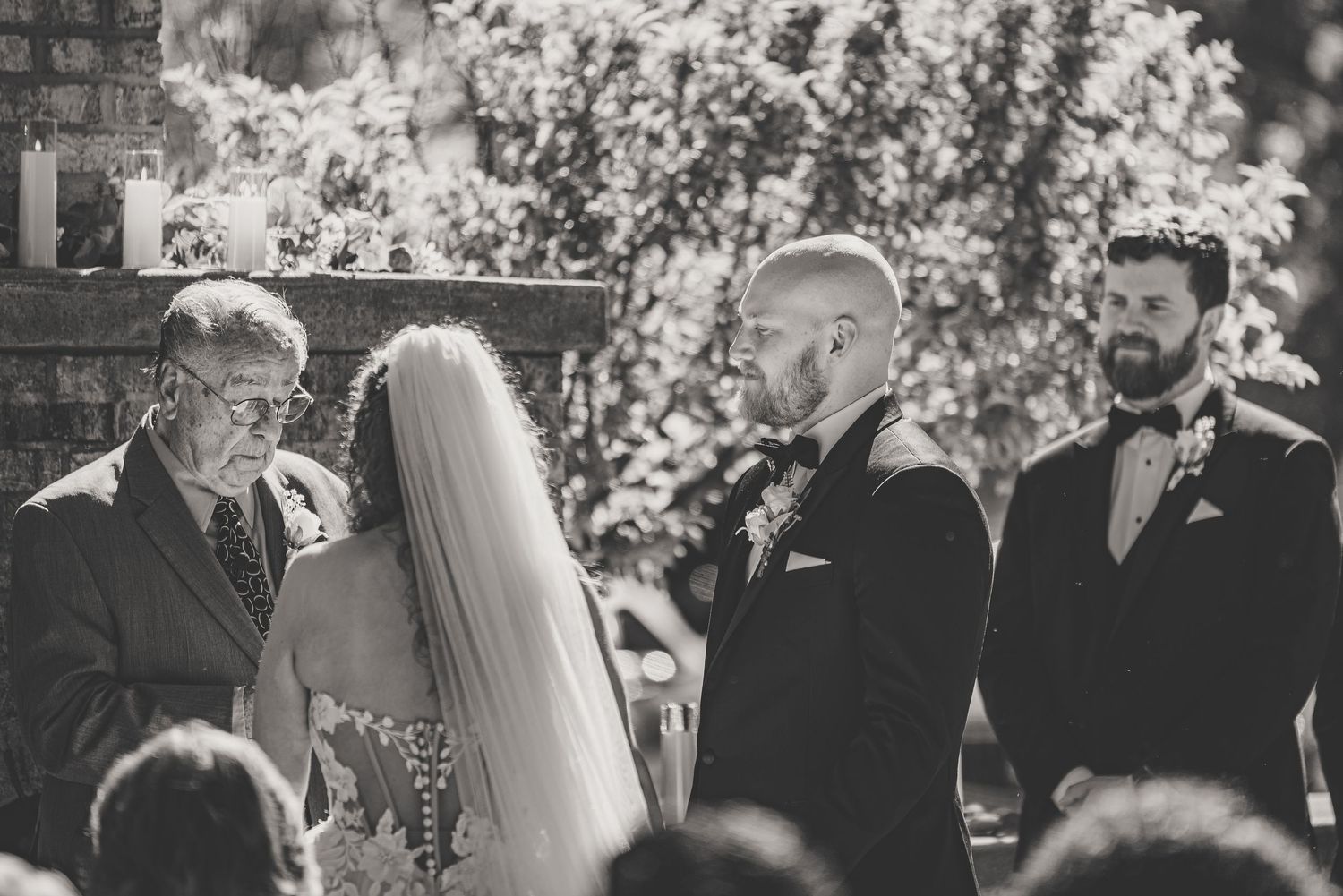 Black and white wedding ceremony photo with officiant, groom, and best man standing at the altar outdoors.
