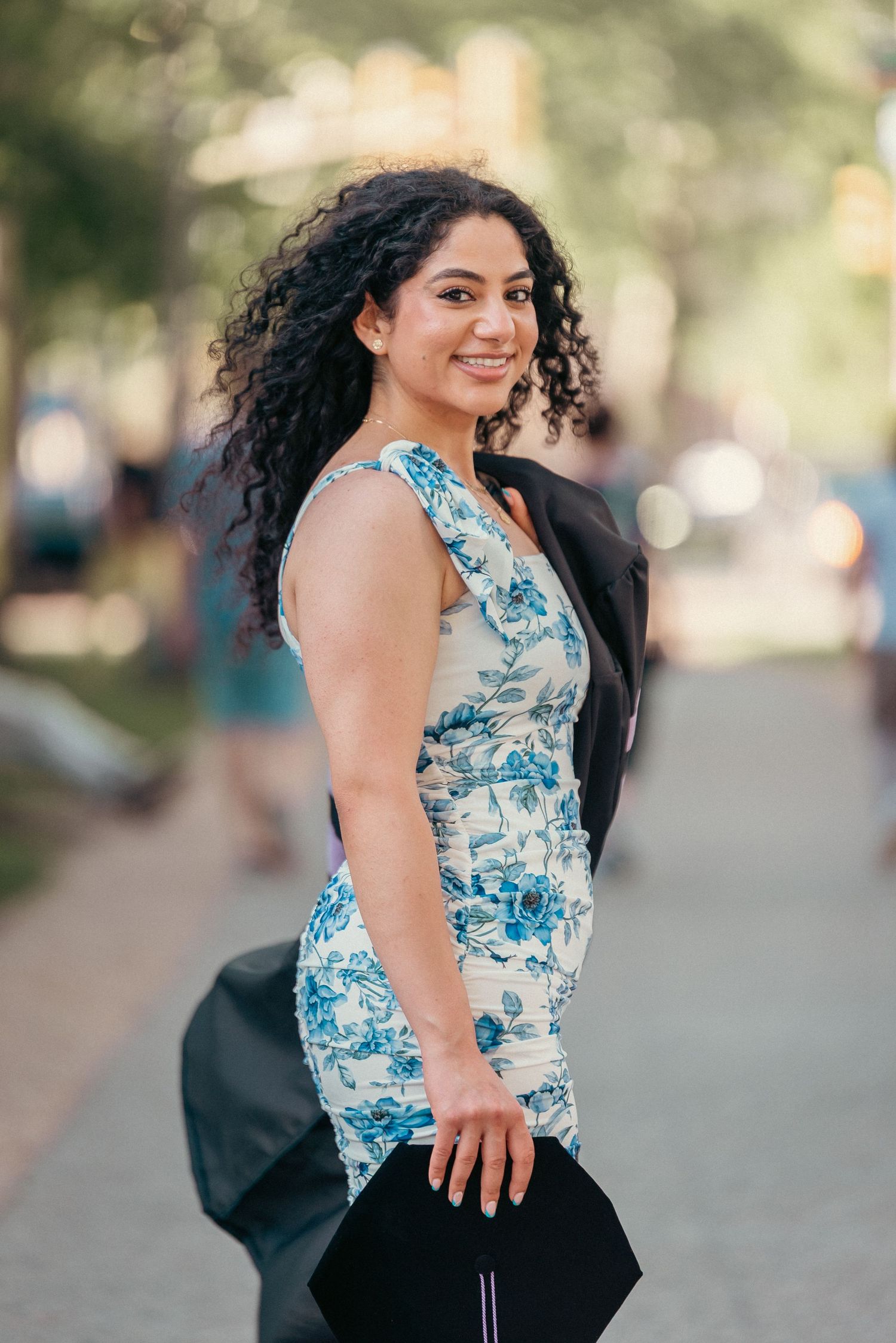 Woman in a blue floral dress smiling over her shoulder outdoors, holding a graduation cap on a tree-lined path.