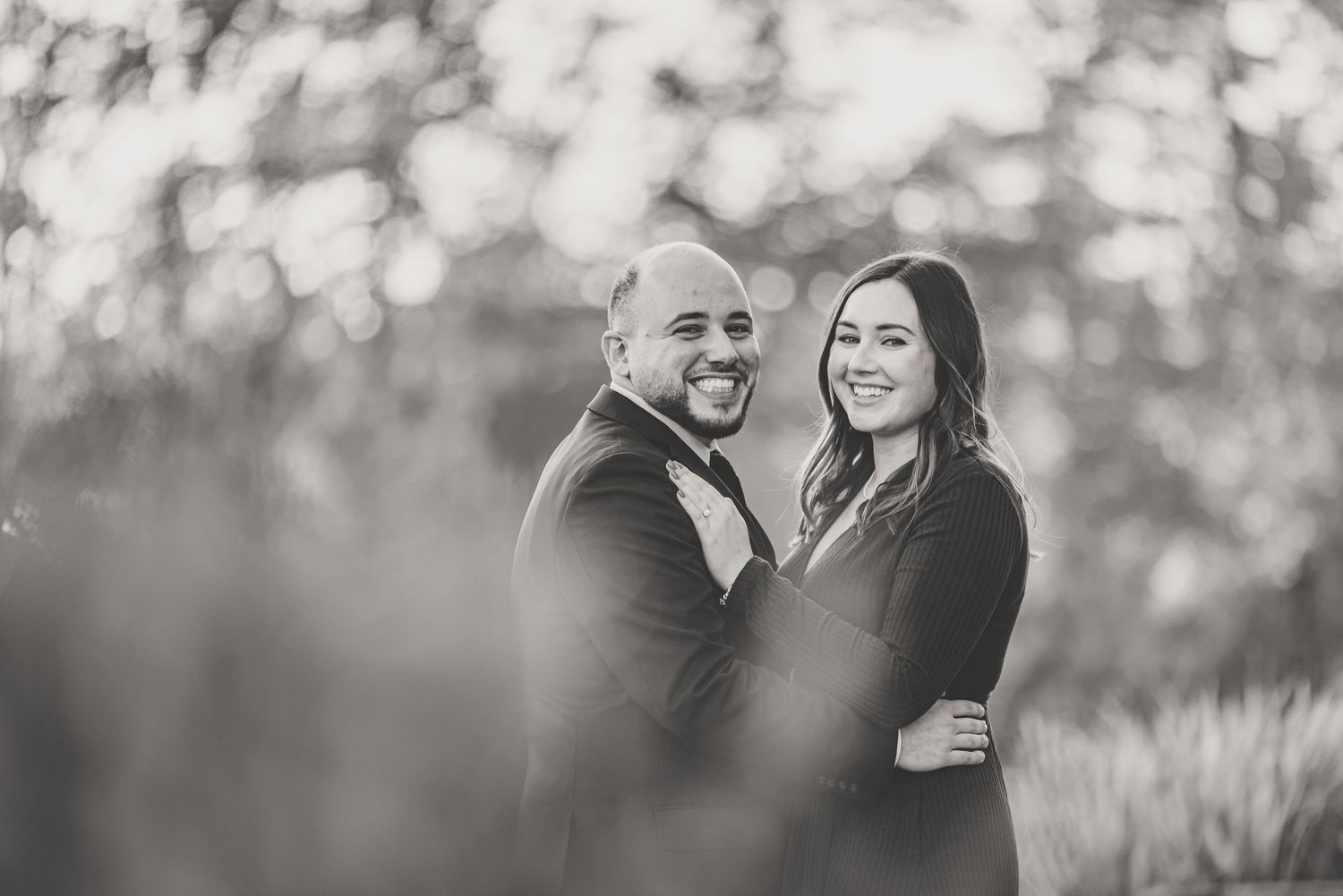 Black and white romantic portrait of engaged couple smiling together in soft bokeh outdoor setting.