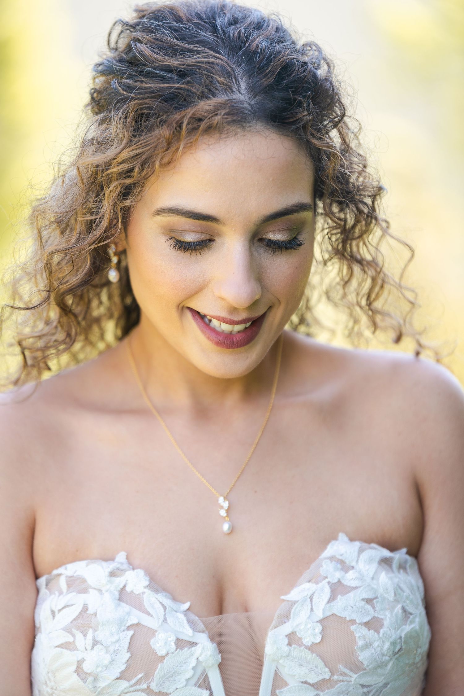 Close-up portrait of smiling bride with curly hair wearing pearl necklace and floral lace gown.
