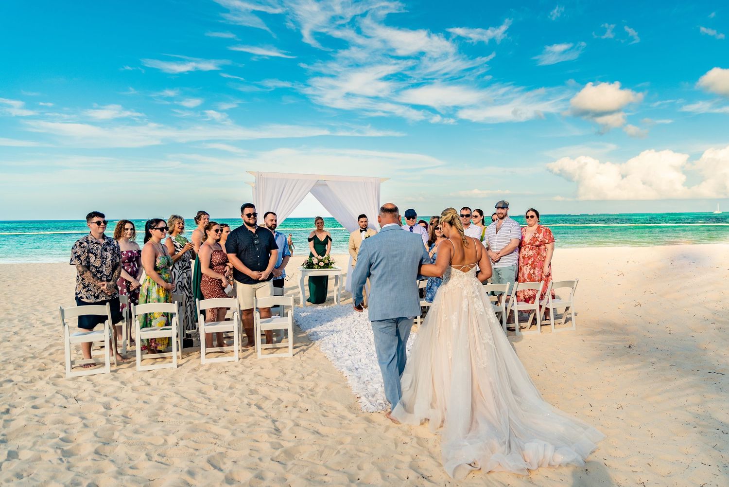 Couple exchanges vows at intimate beach wedding ceremony with turquoise ocean backdrop and white chairs.