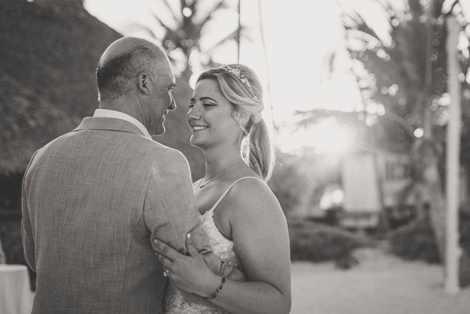Bride smiles warmly during dance with partner at golden hour tropical beach wedding celebration.
