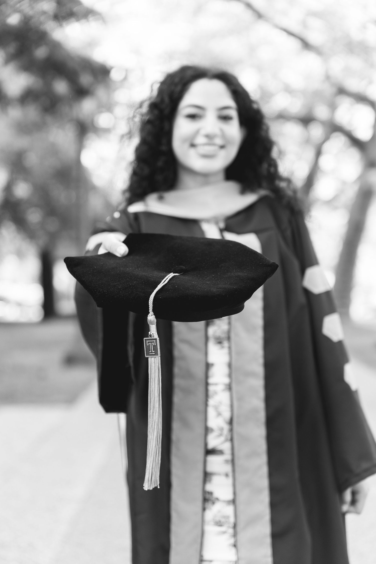Smiling graduate in black cap and gown holding mortarboard toward camera in black and white photo.
