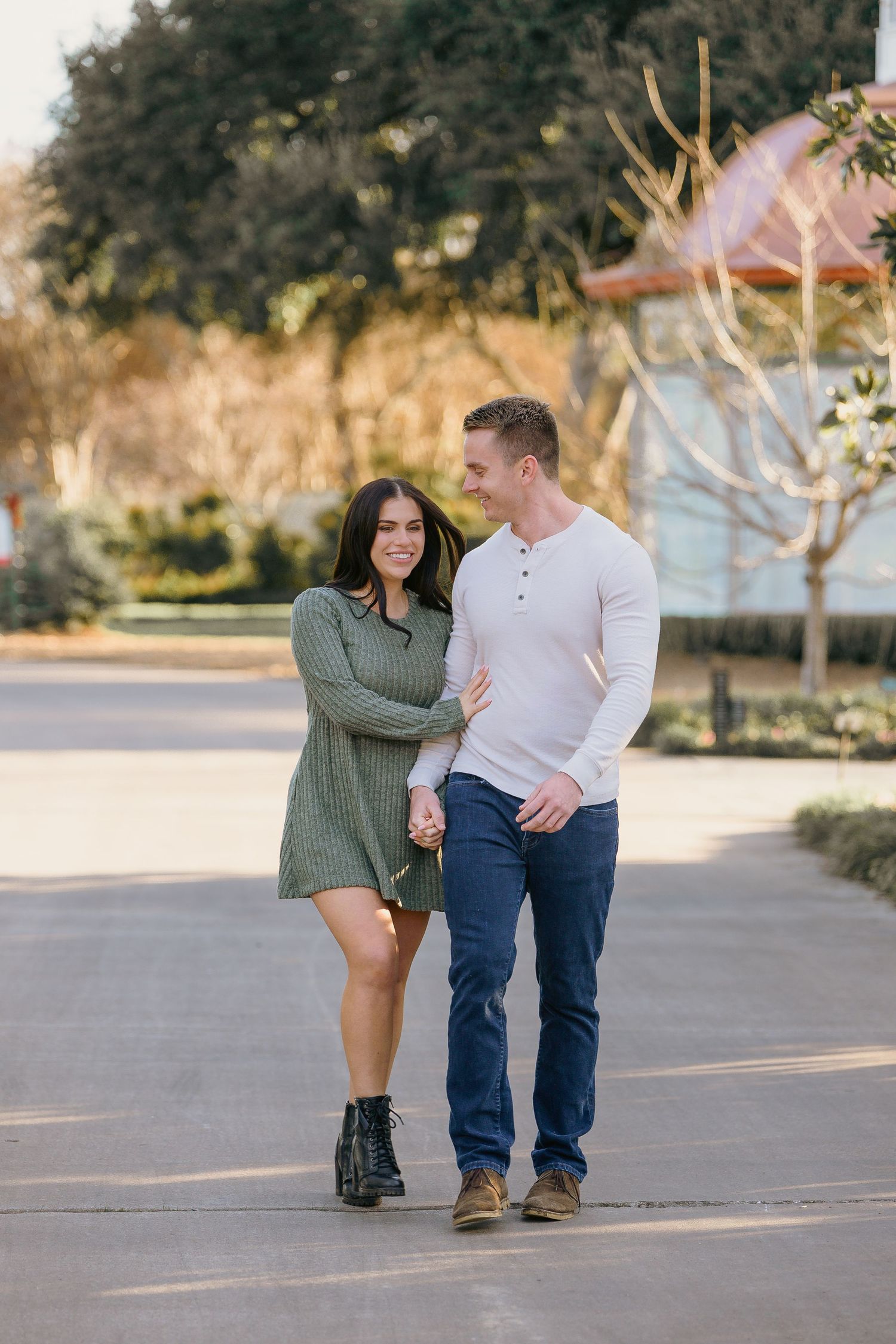 A couple walks hand in hand along a sunlit path, smiling at each other in a park setting.