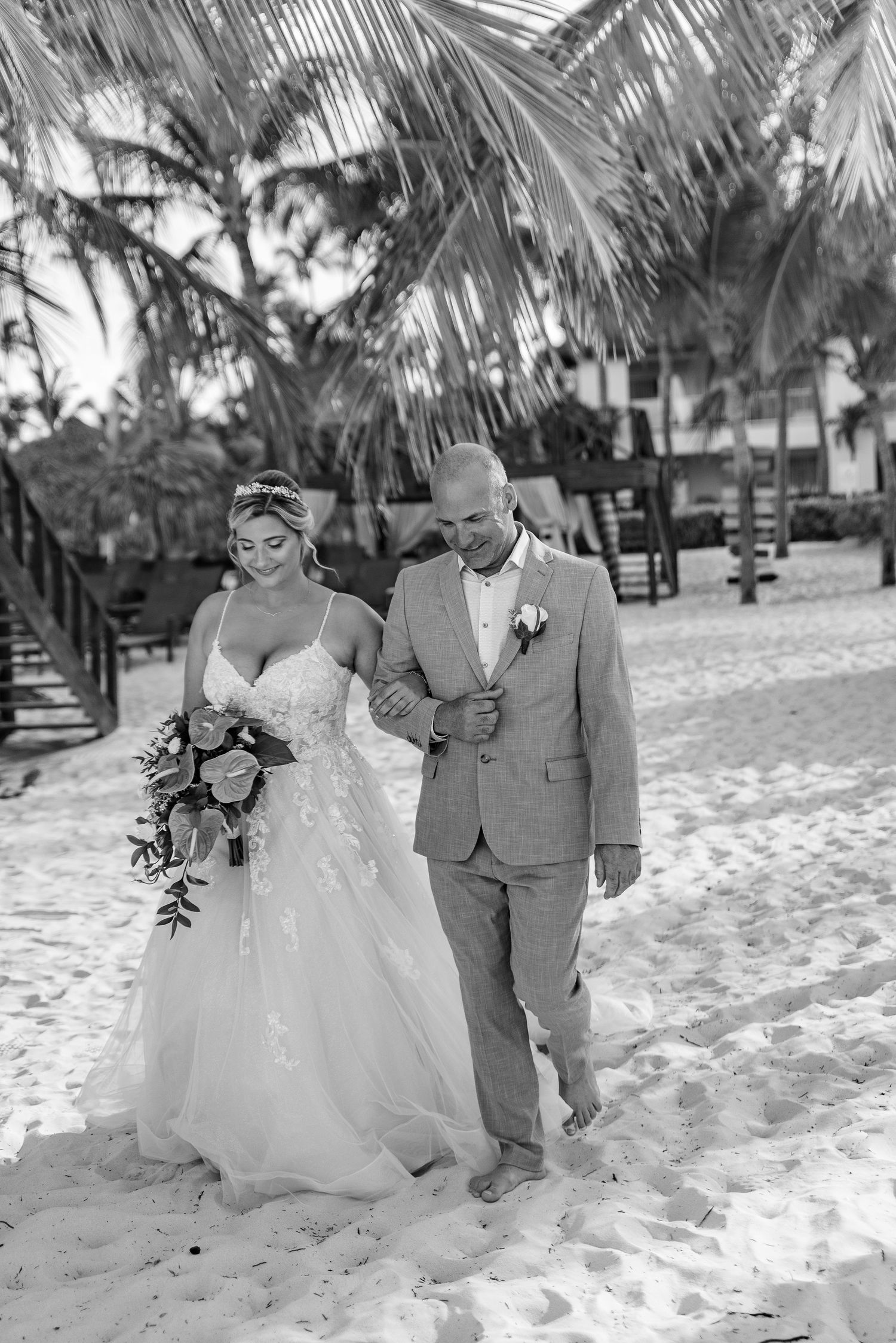 Black and white photo of bride in lace gown walking with escort along a sandy beach lined with palms.