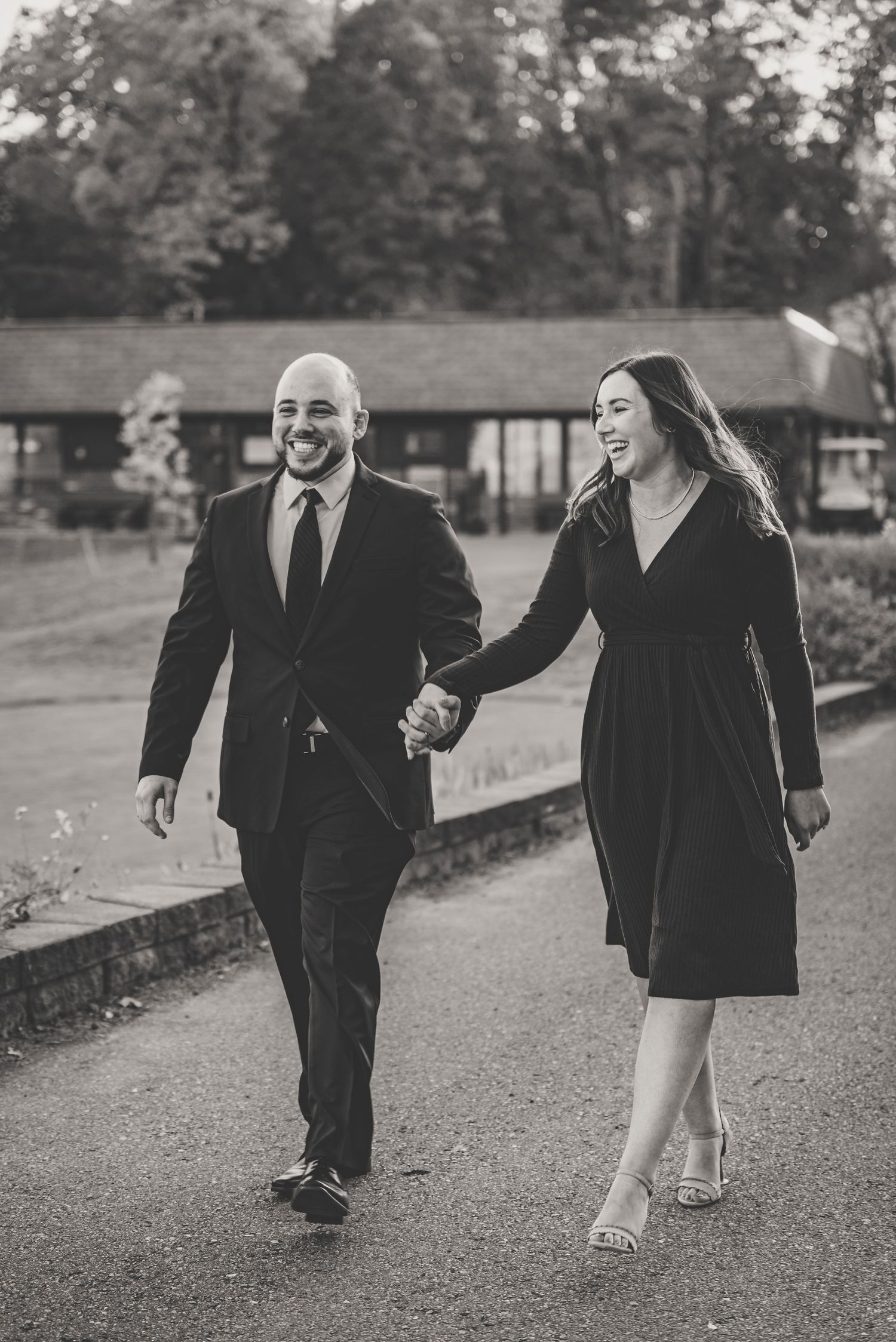 Smiling couple holding hands walking outdoors in black and white engagement photo at venue.
