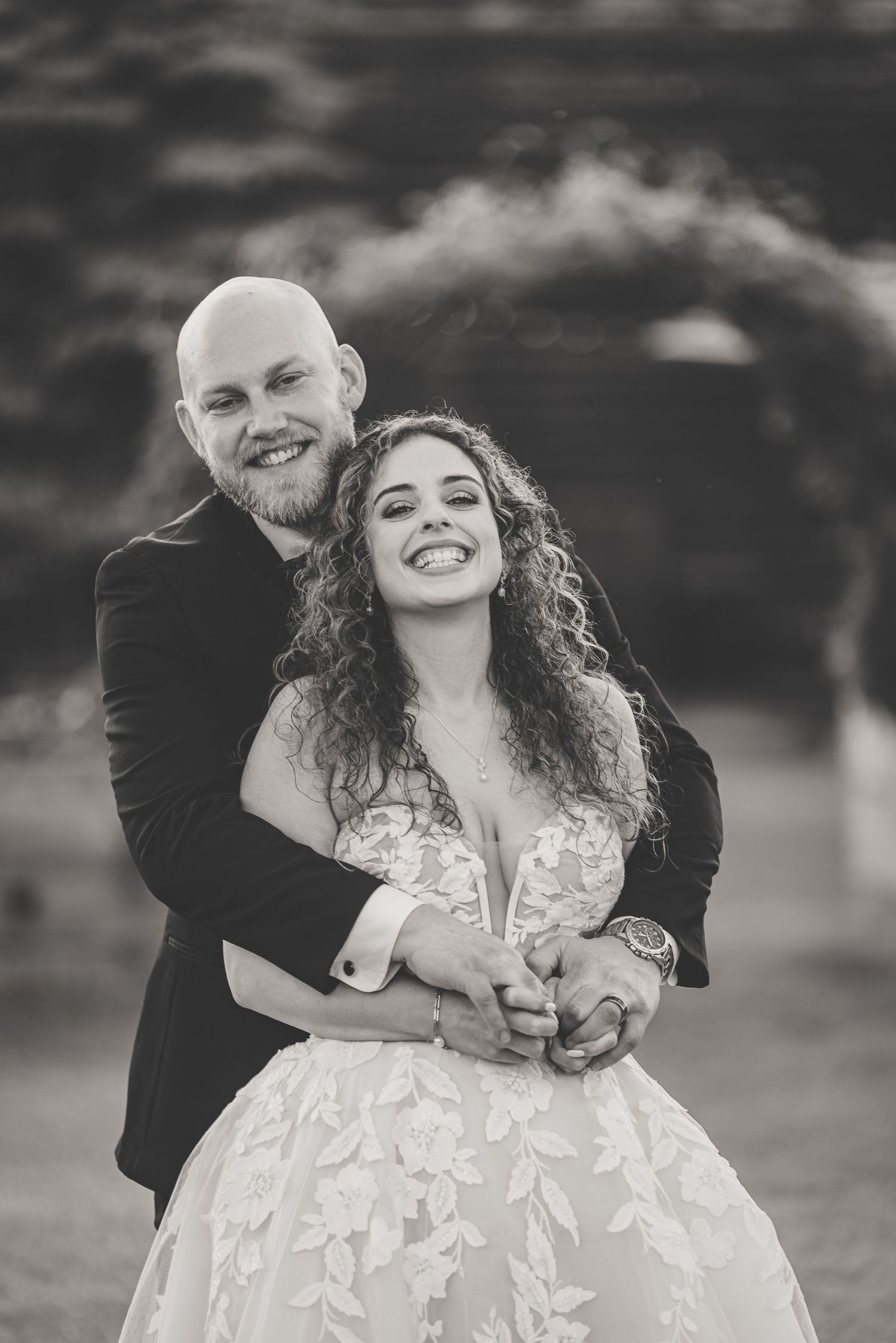 Black and white photo of groom embracing bride from behind, both smiling joyfully in outdoor wedding setting.
