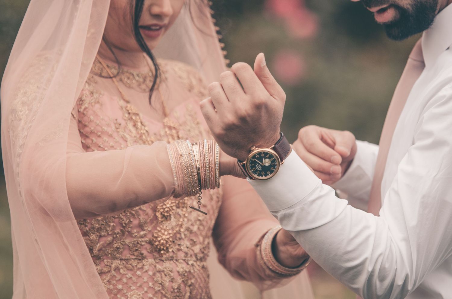 Bride in pink lehenga with gold jewelry adjusting groom's watch cufflinks during Indian wedding ceremony.