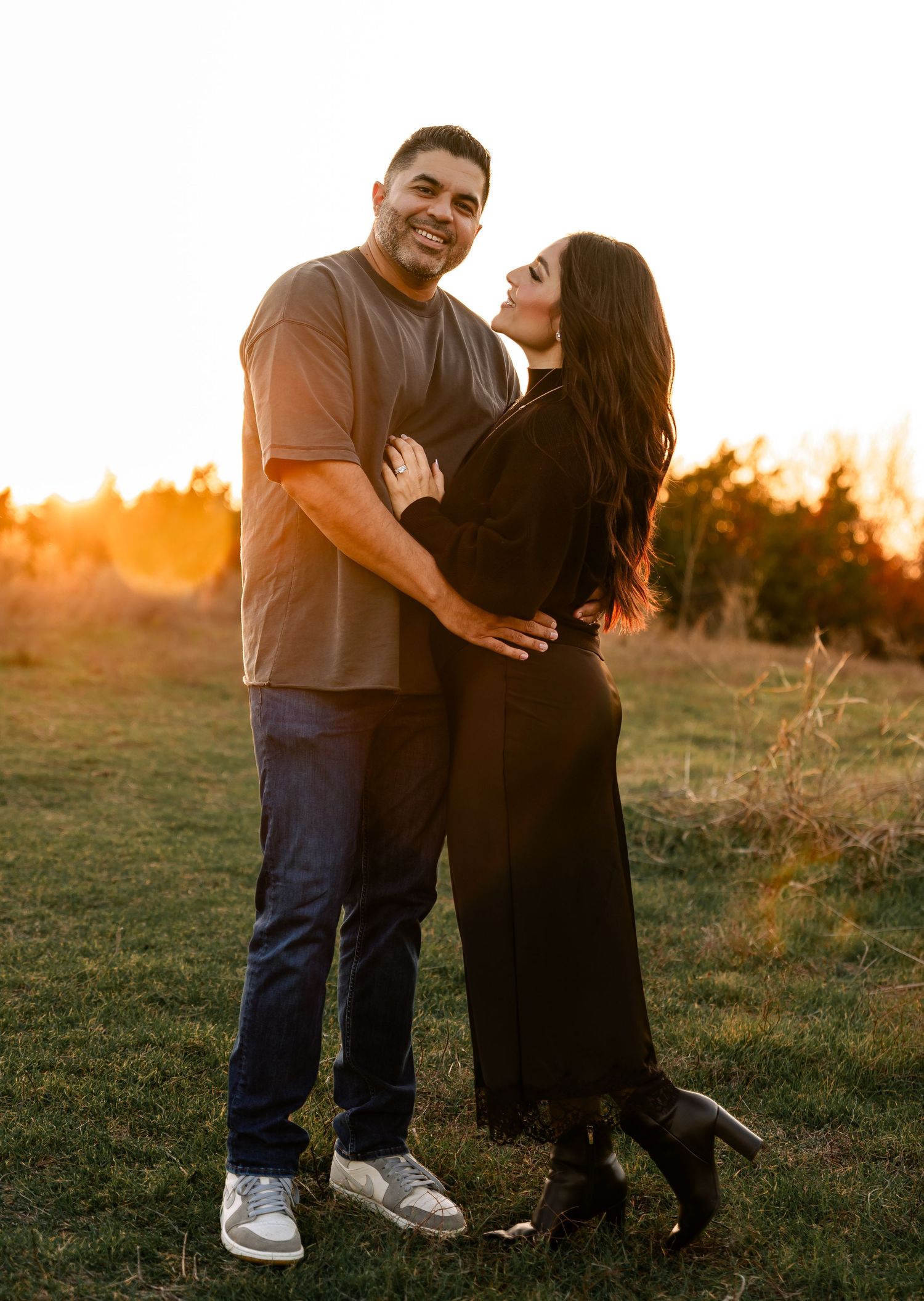 A happy couple poses together in a golden sunset field, the woman looking up joyfully at her partner.