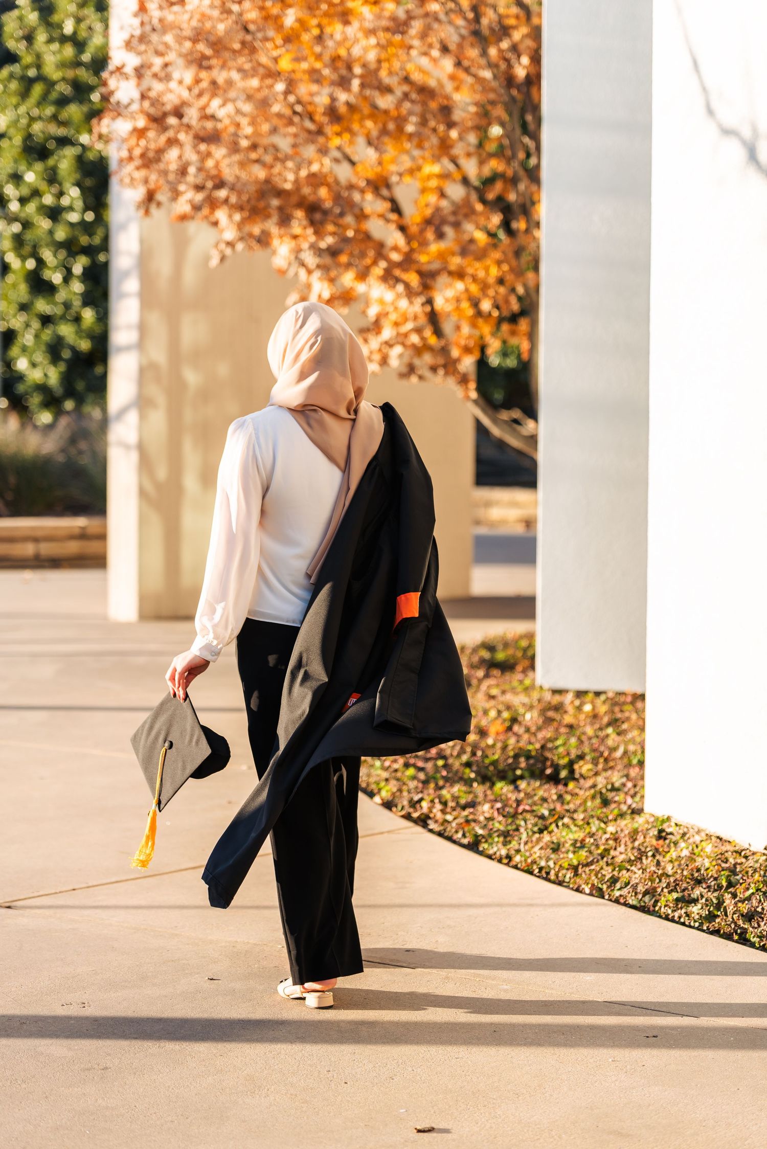 A hijab-wearing graduate walks outdoors carrying her cap and gown in autumn sunlight.