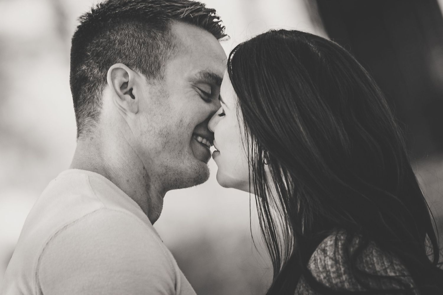 Black and white close-up of a smiling couple leaning in for a kiss with soft bokeh background.
