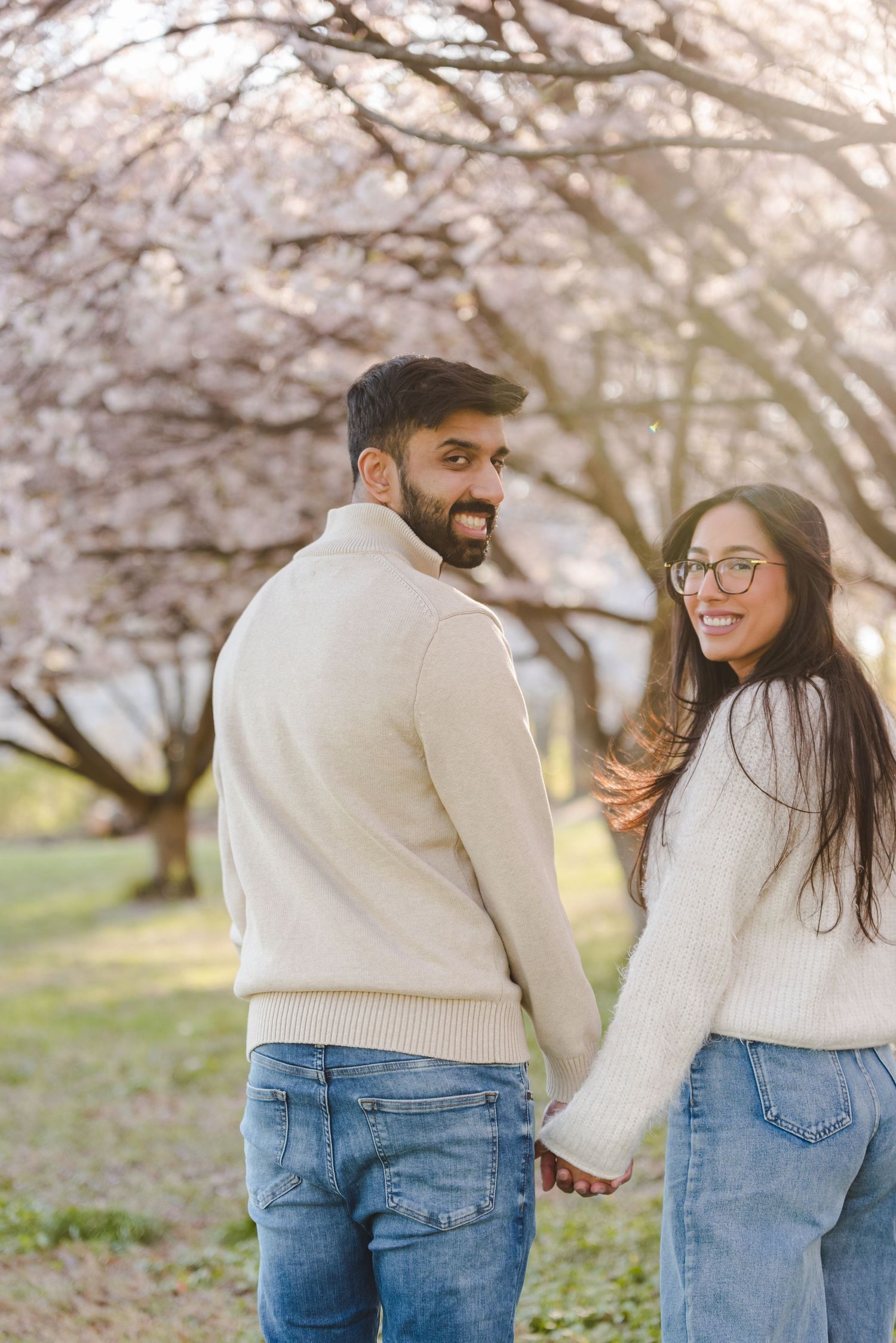 Smiling couple holding hands and looking back at camera under pink cherry blossom trees in matching cream sweaters.