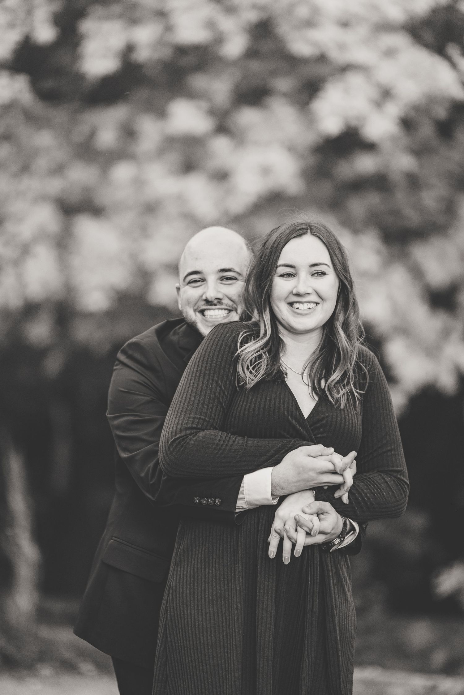 Black and white portrait of smiling couple embracing with man hugging woman from behind outdoors.