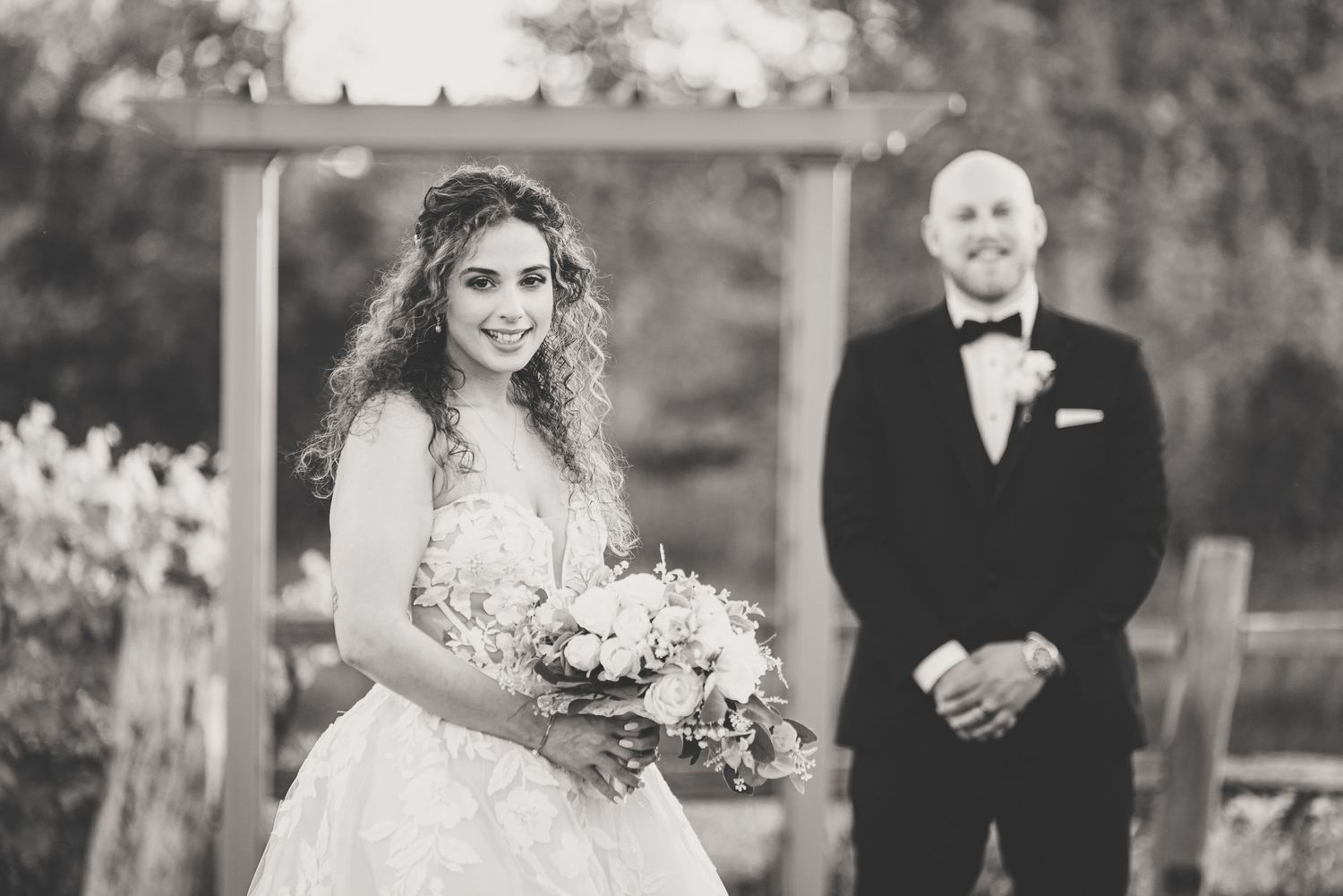 Black and white photo of smiling bride holding bouquet in lace gown with groom standing behind her outdoors.