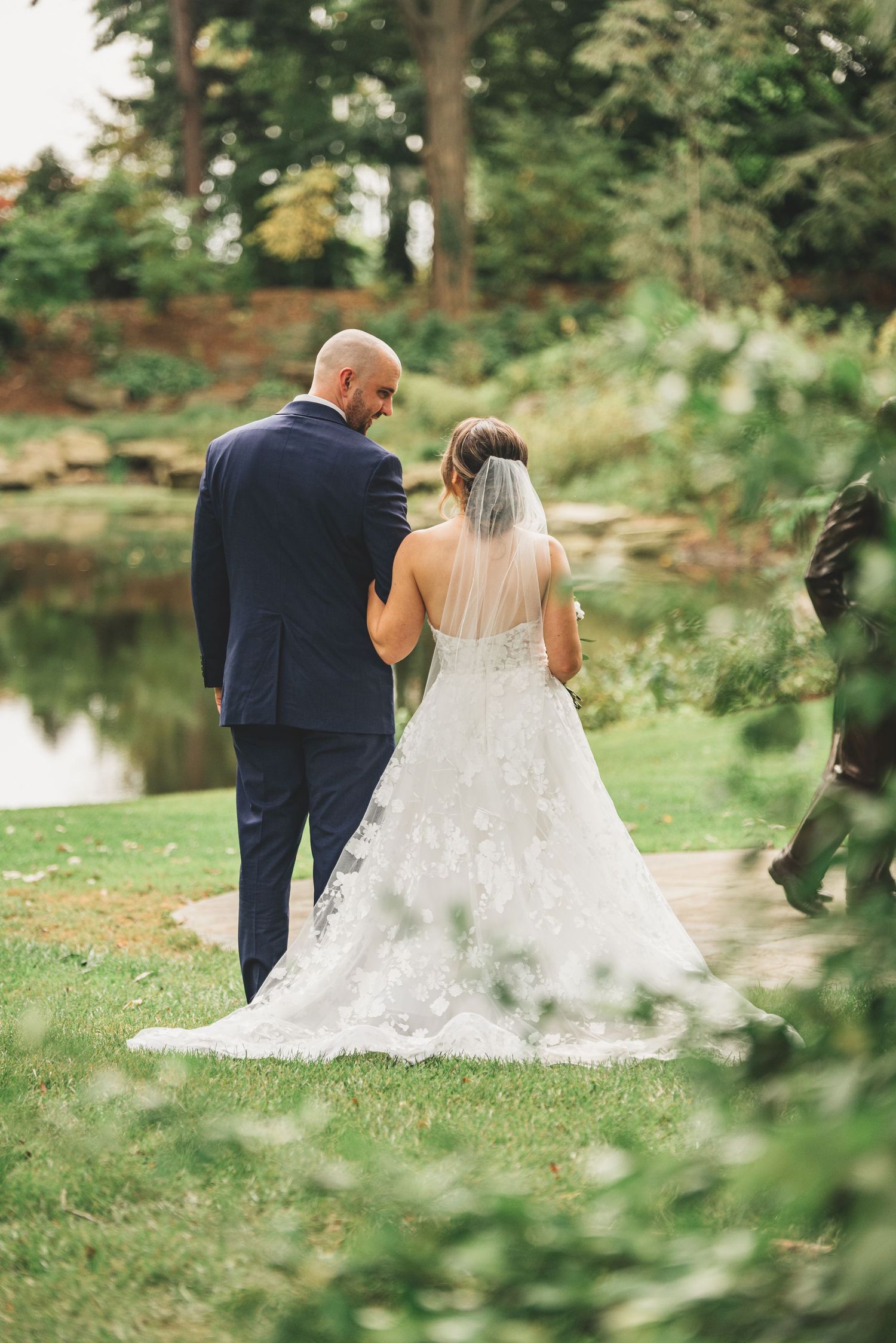 Bride and groom in navy suit walking through scenic garden near pond after wedding.