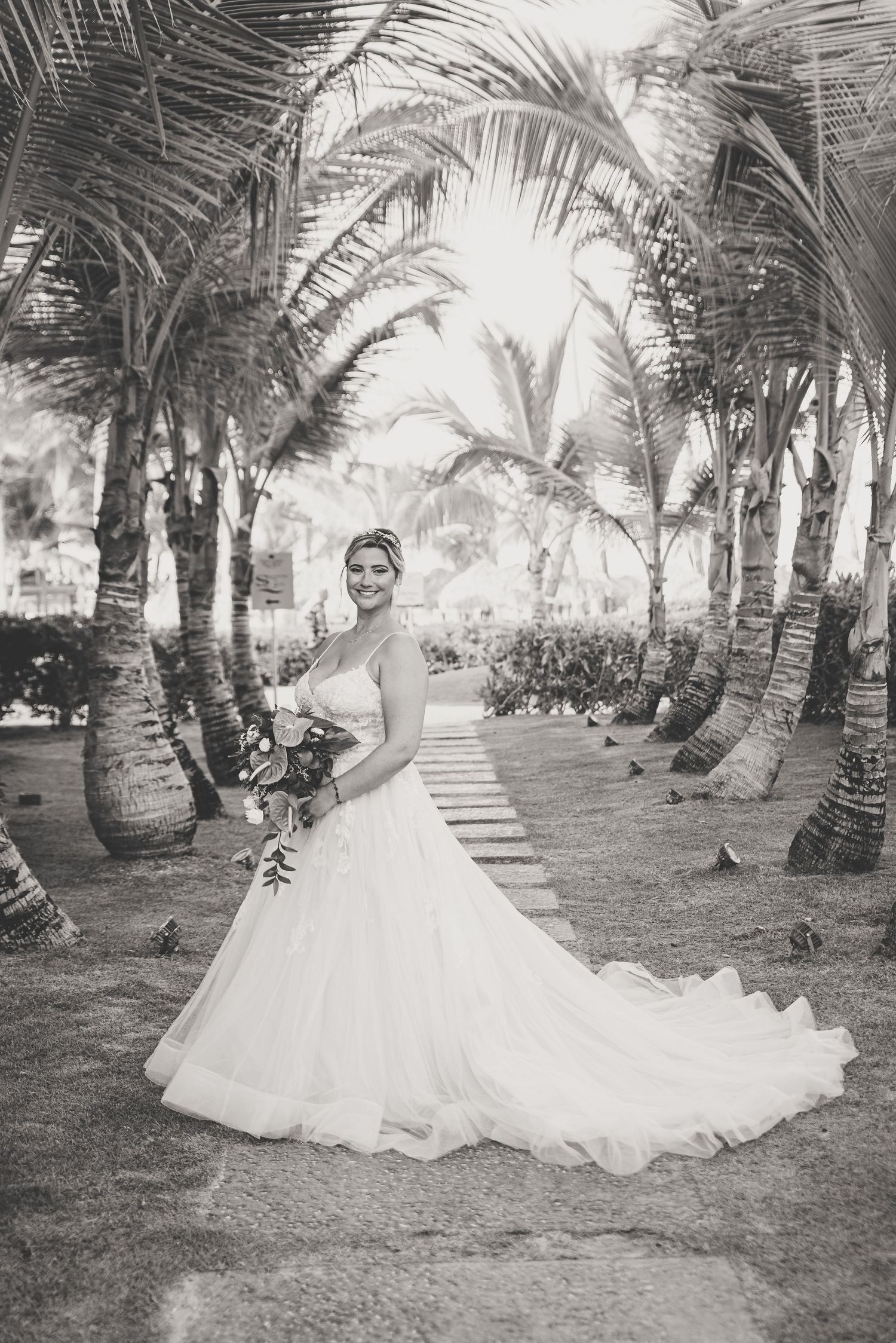 Bride in elegant ball gown poses among tropical palm trees in classic black and white portrait.