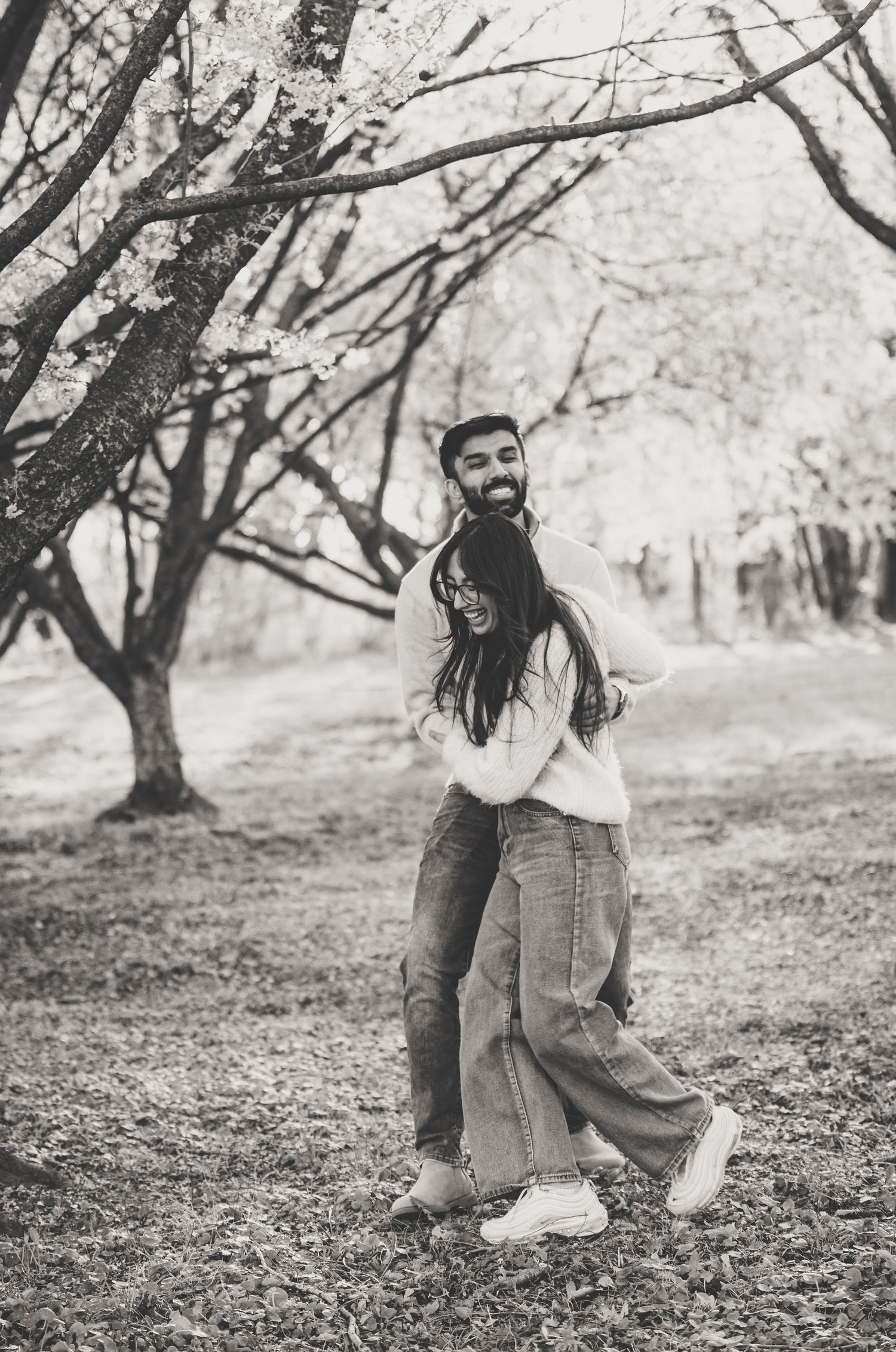 Black and white photo of a couple embracing and laughing together under bare trees in a park.