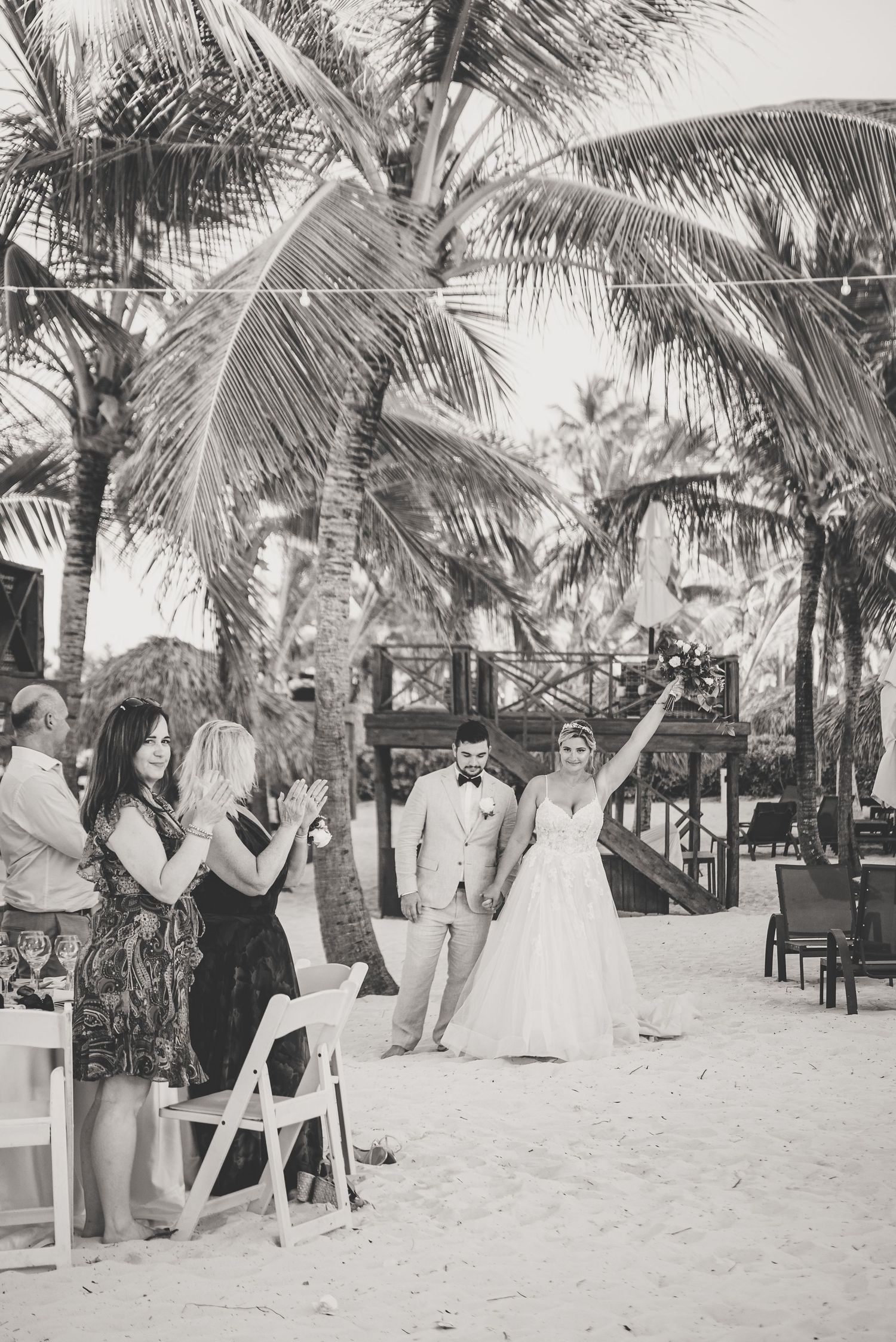 Bride raises arm in celebration as couple walks through guests at tropical beach wedding reception.