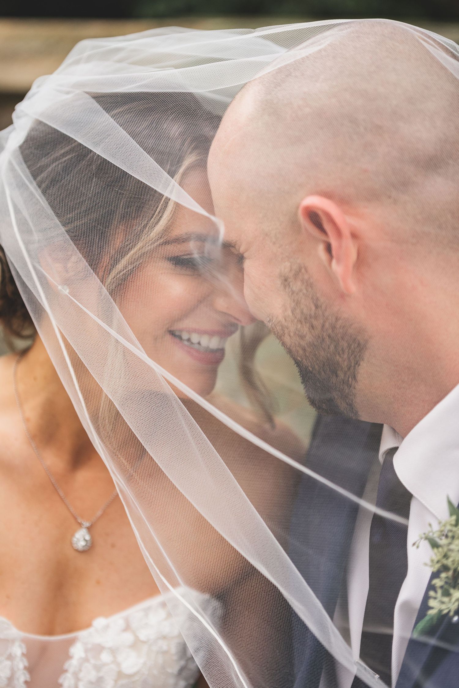 Bride and groom sharing intimate moment under flowing wedding veil, touching foreheads and smiling.