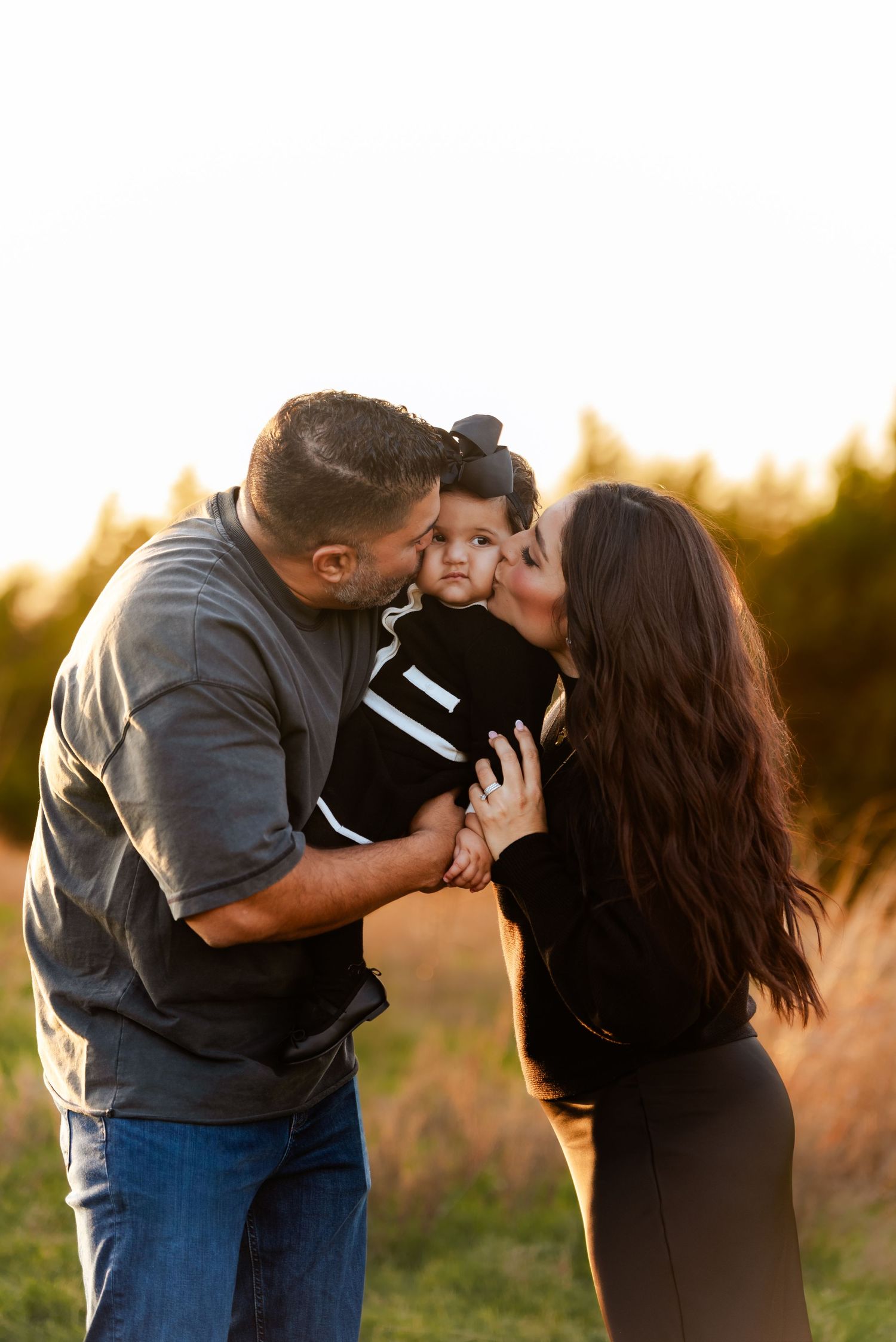 Two parents lovingly kiss their baby on the cheeks during an outdoor golden hour family portrait session.