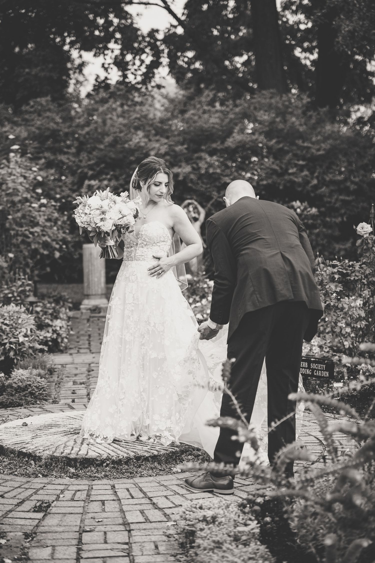 Bride in lace gown holding bouquet assisted by man in garden ceremony.