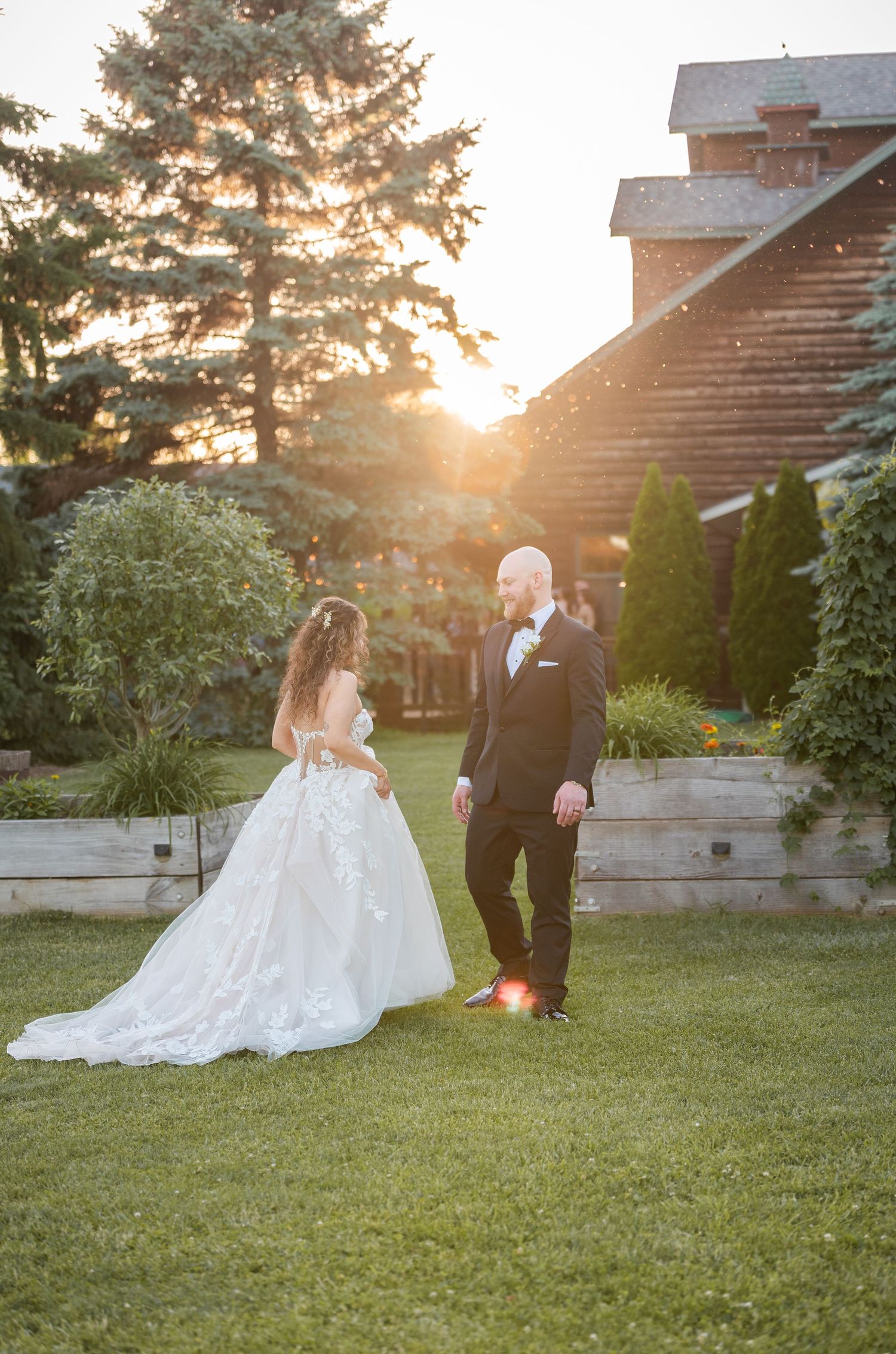 Bride and groom share a golden sunset moment on lush garden grounds near a rustic barn venue.