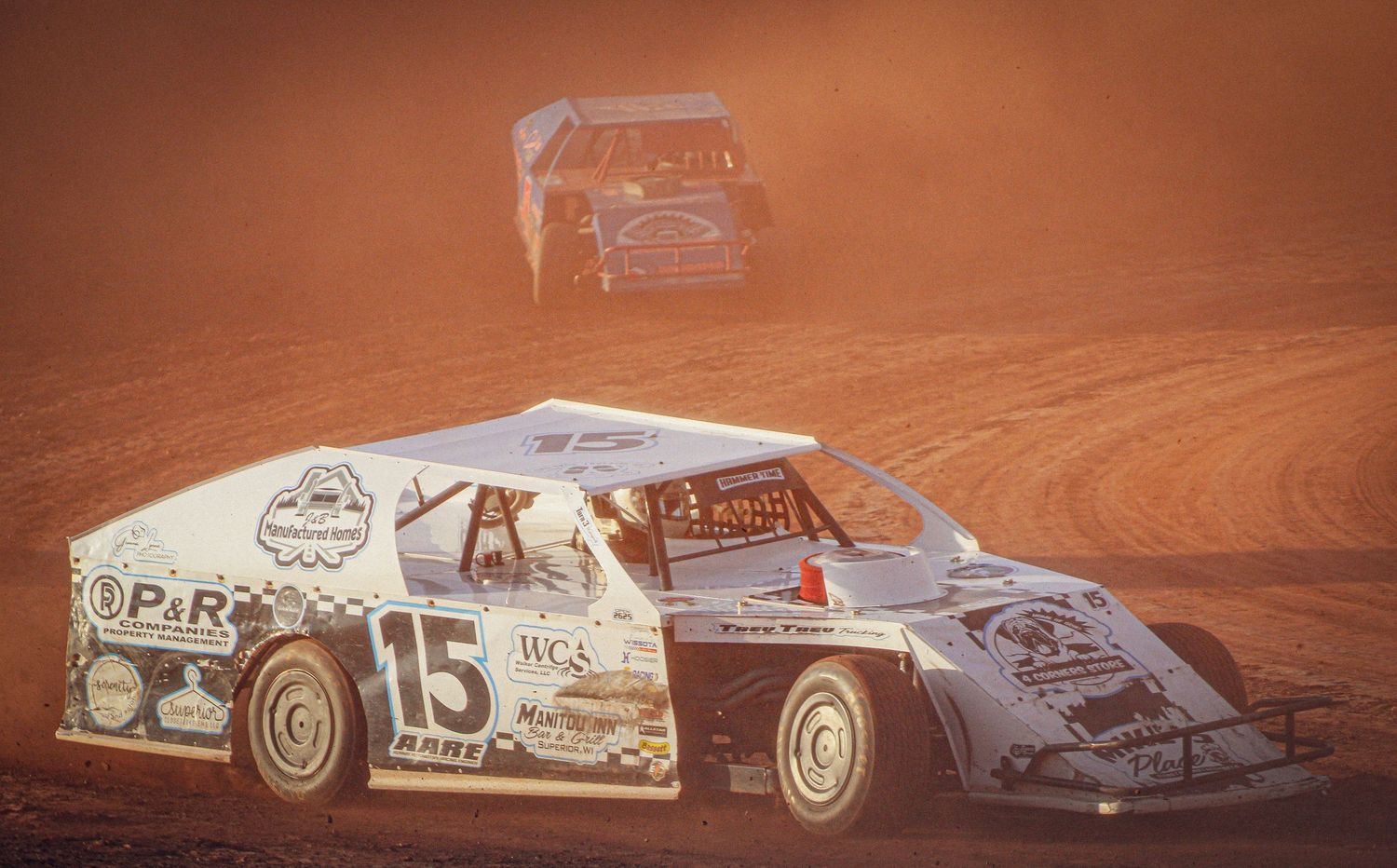 Two dirt track race cars, including car number 15, compete on a dusty oval racing circuit.