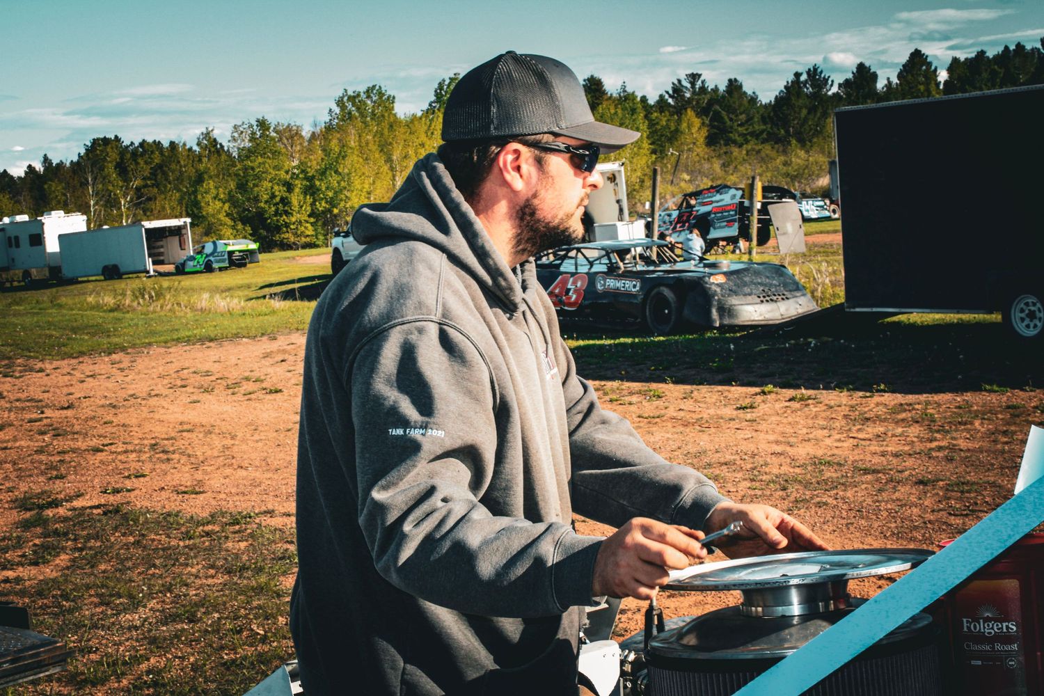 Person working on equipment outdoors at a racing event with trees in background.