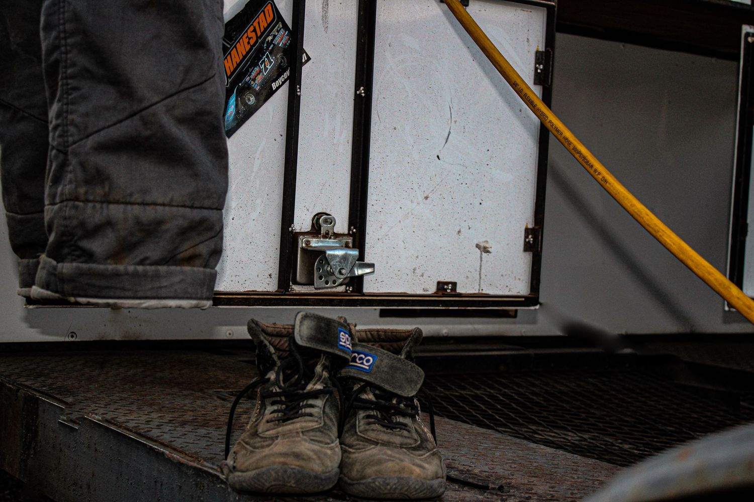 Work boots and jeans sit on a metal grate near a white door with a yellow safety bar.
