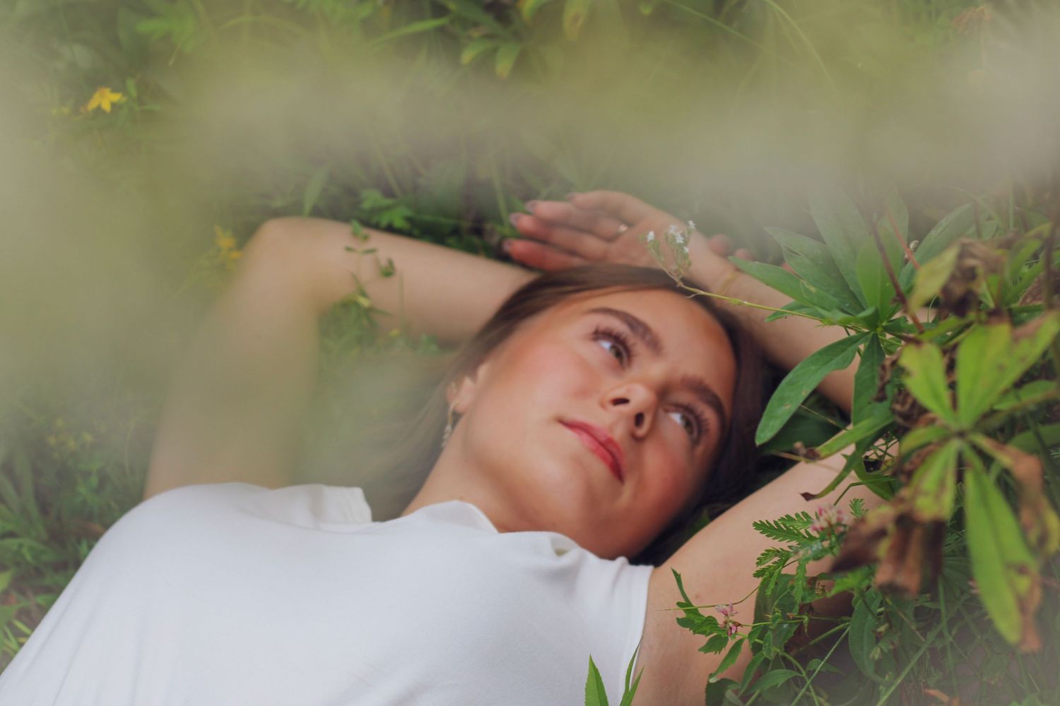 Woman in white top lying among lush green plants and ferns, gazing upward in a serene outdoor setting.