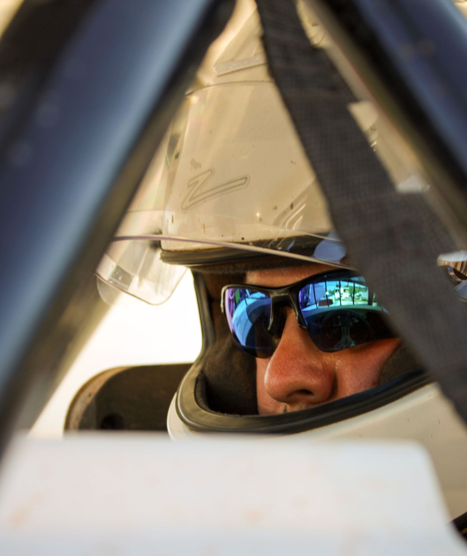 View through race car frame showing driver wearing reflective sunglasses in cockpit.
