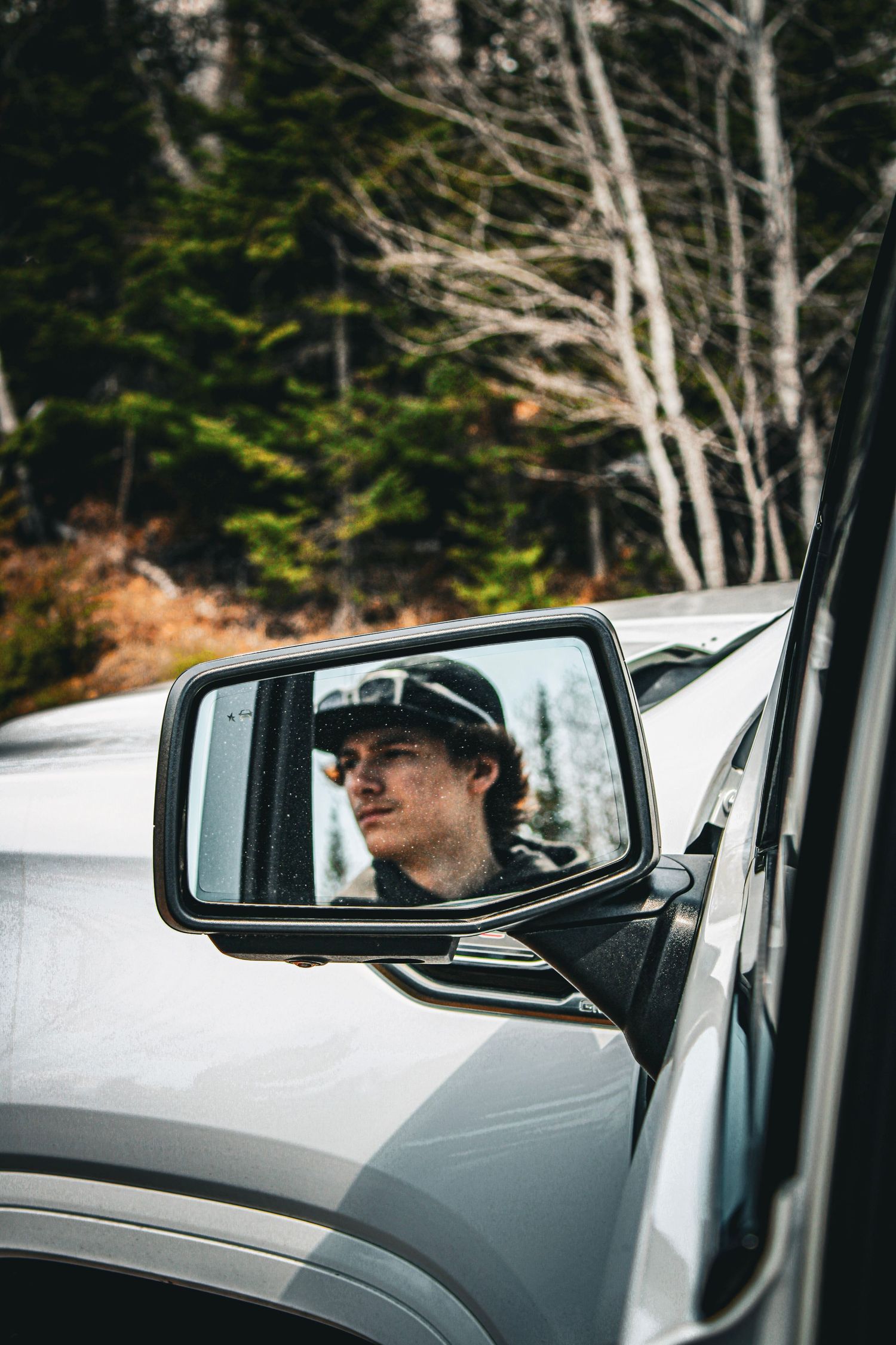 Person reflected in car side mirror wearing a cap, surrounded by forest trees in the background.