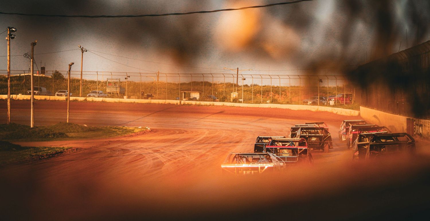 Dirt track racing cars compete under bright lights on a red clay oval speedway at night.