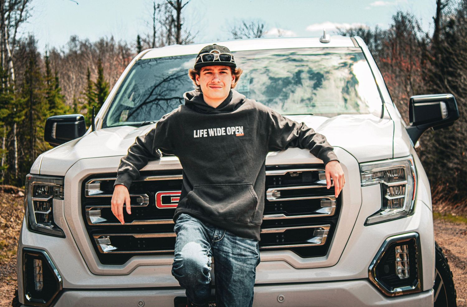 Young man in a black hoodie leaning on a white GMC truck outdoors in a wooded area.