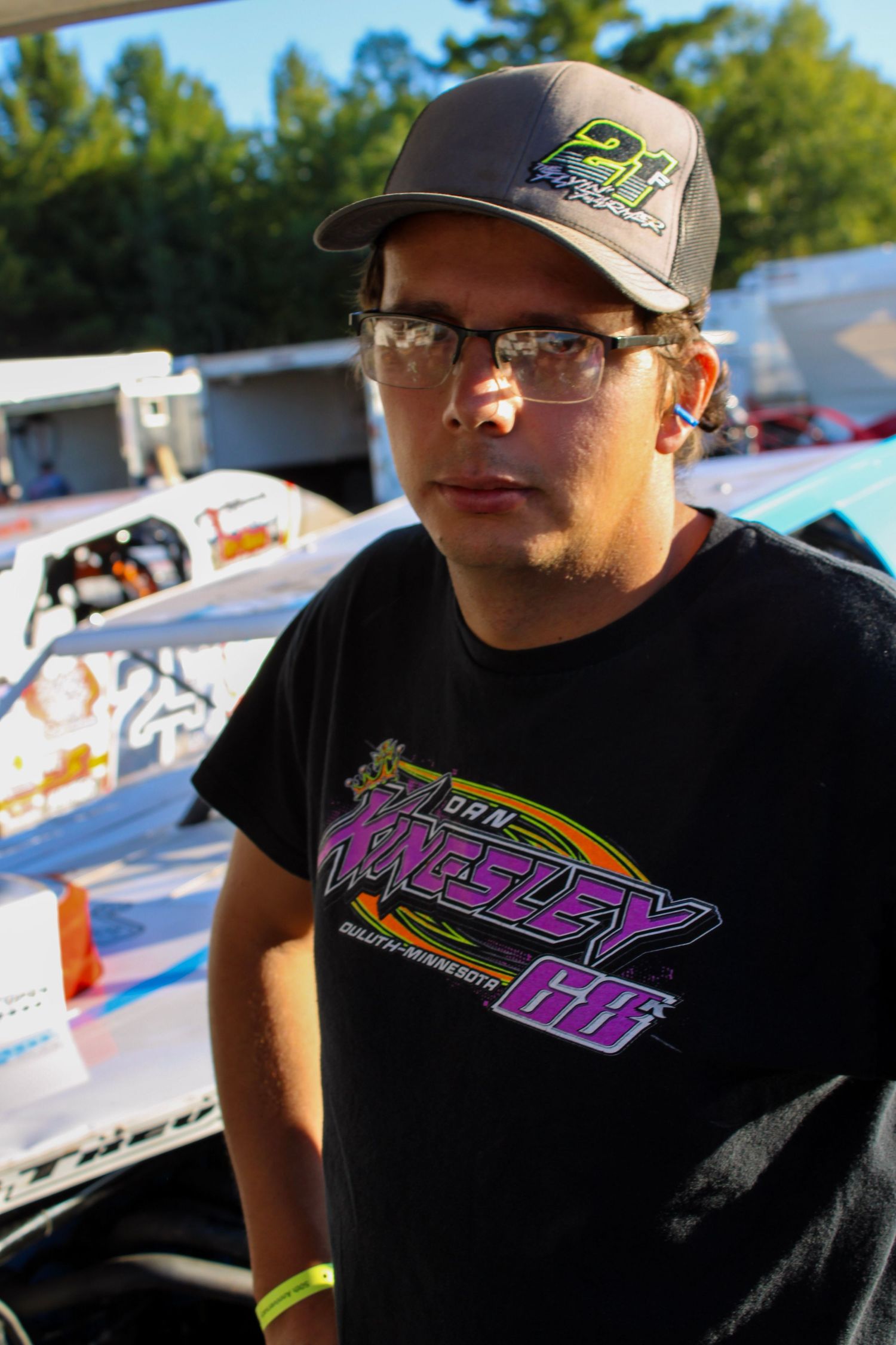 Racing team member wearing branded cap and t-shirt stands by pit area.