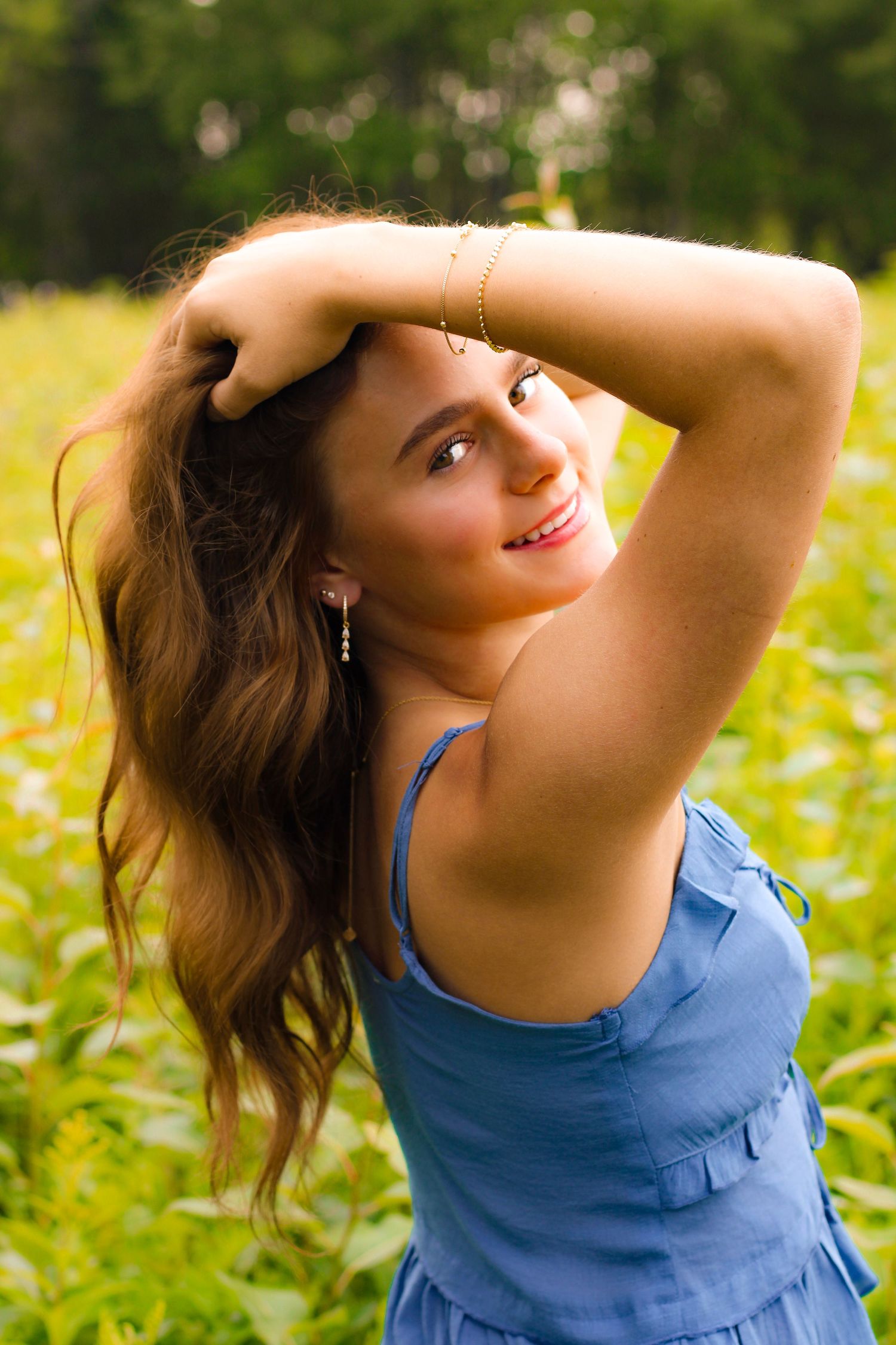 Young woman in blue dress posing outdoors in green field, smiling with hand raised to her long brown hair.