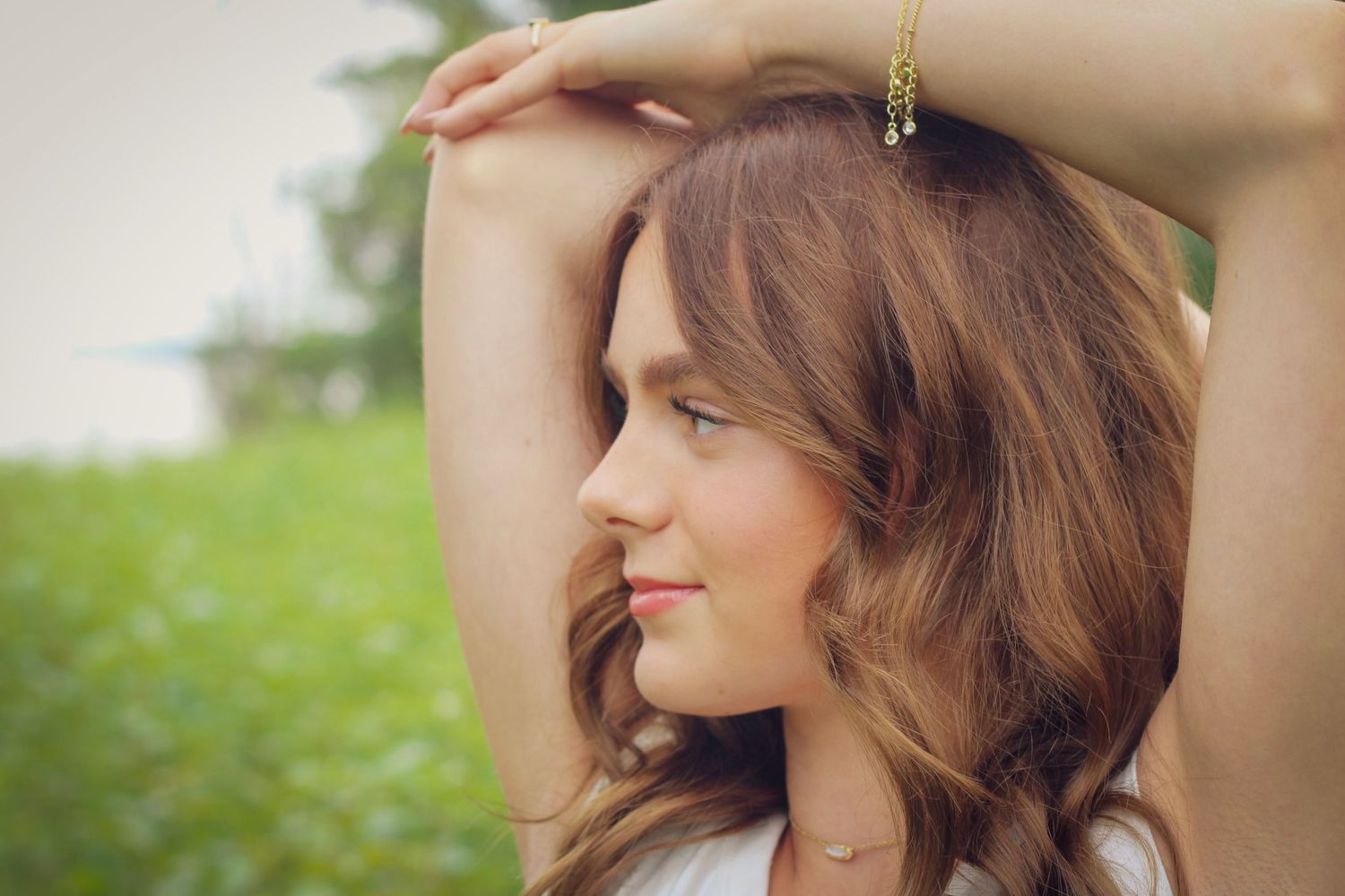 Young woman with brown wavy hair poses outdoors, arms raised gracefully, wearing a white top and gold bracelet.