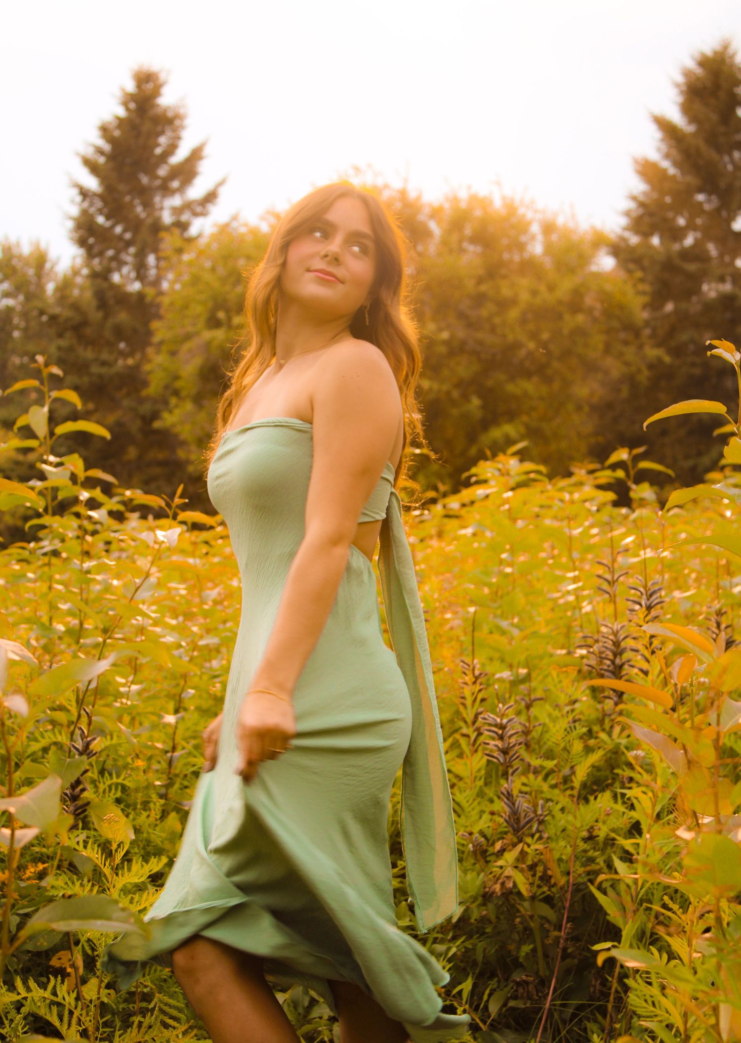 Woman in sage green midi dress standing in a sunlit wildflower field, looking over her shoulder.