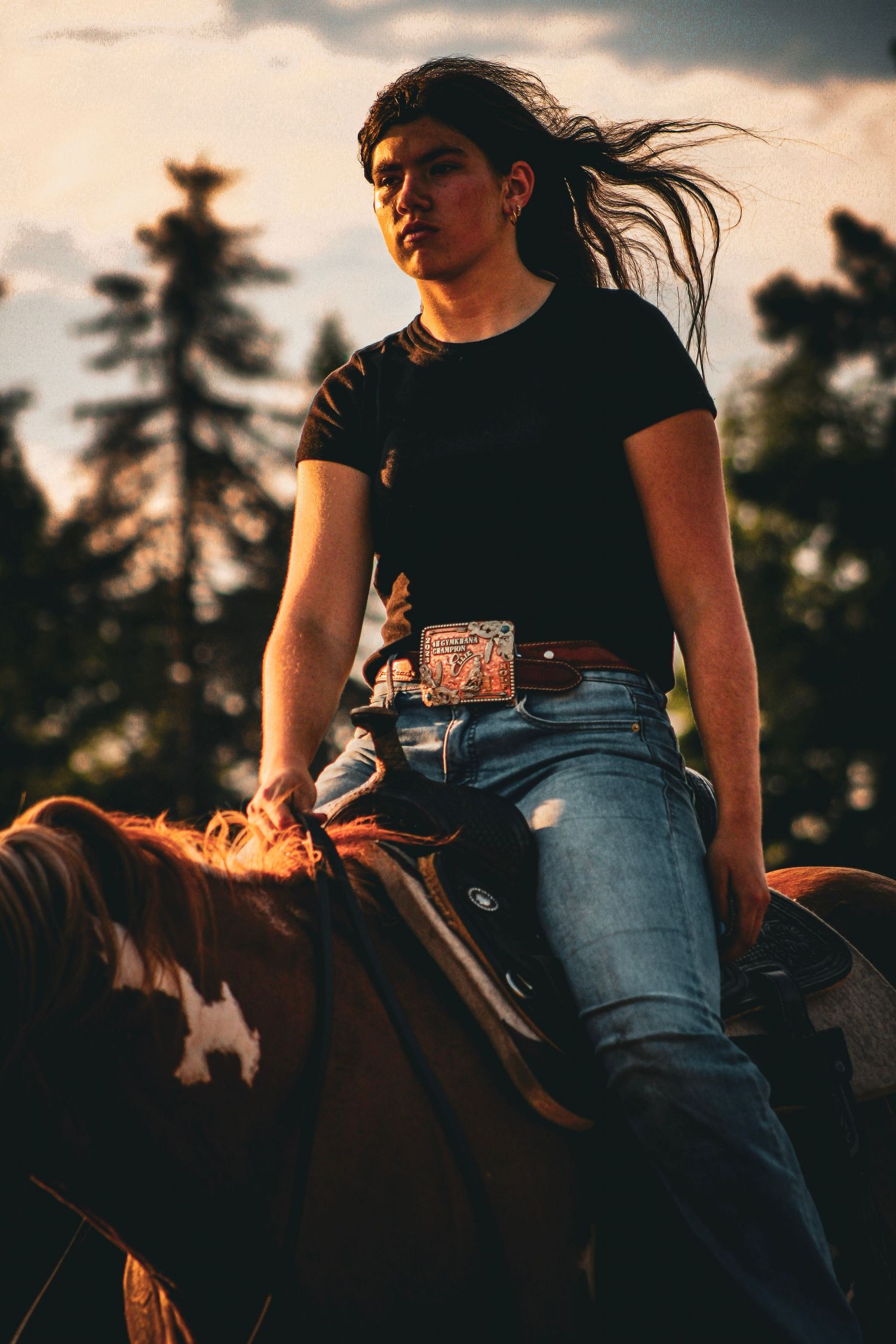 Young rider on horseback at sunset, wearing black tee, jeans, and decorative belt, with hair blowing in wind.